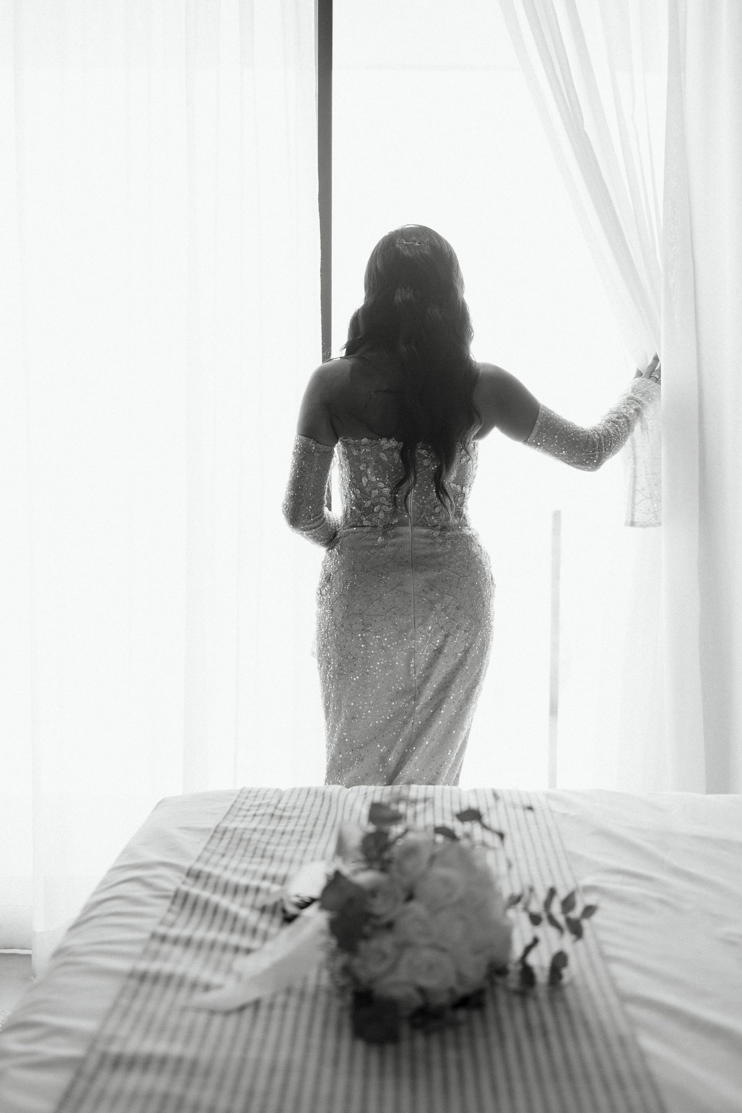 Black and white photo of bride in lace gown standing at window with bouquet on bed in foreground.