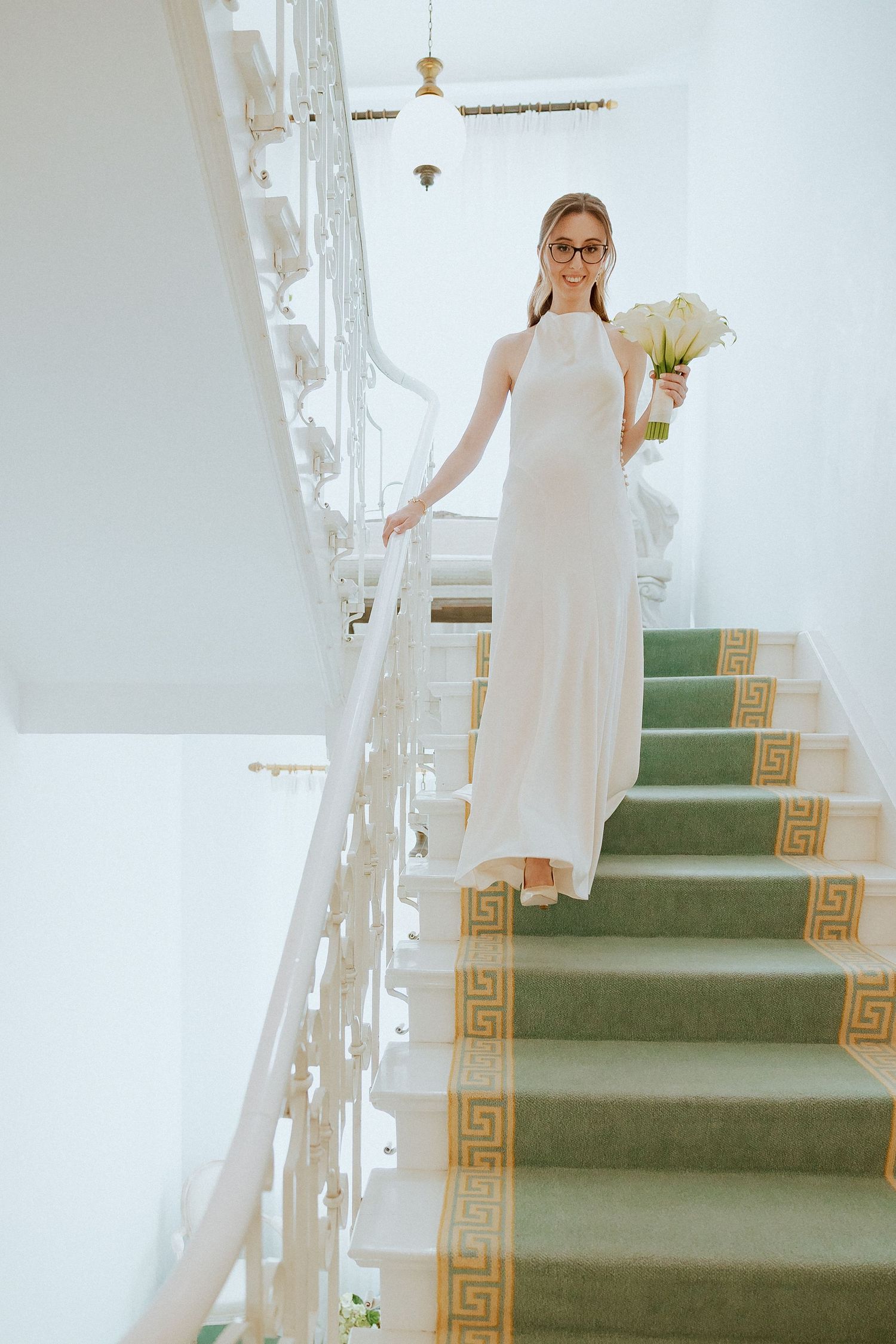 Elegant bride in white gown and glasses descending ornate staircase holding white calla lily bouquet.