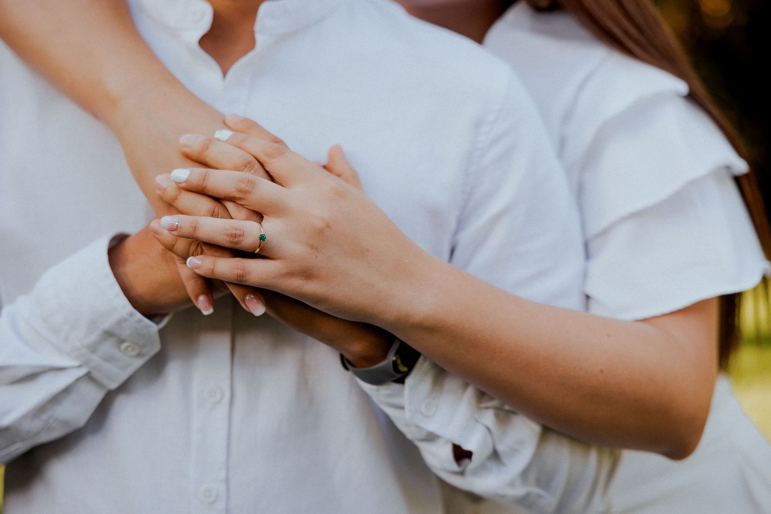Closeup of hands wearing wedding rings and holding each other against white clothing in natural light.