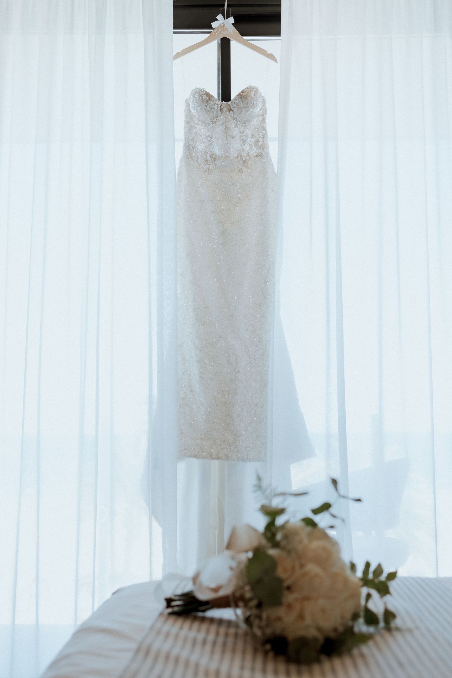 Elegant strapless lace wedding dress hanging on a hanger against sheer white curtains with a bridal bouquet below.