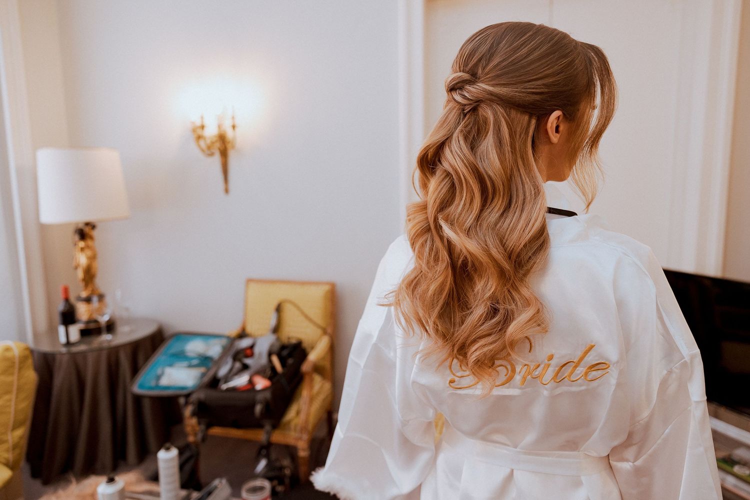 Bride in white satin robe with gold script, showcasing elegant half-up wavy hairstyle in a luxury hotel room.