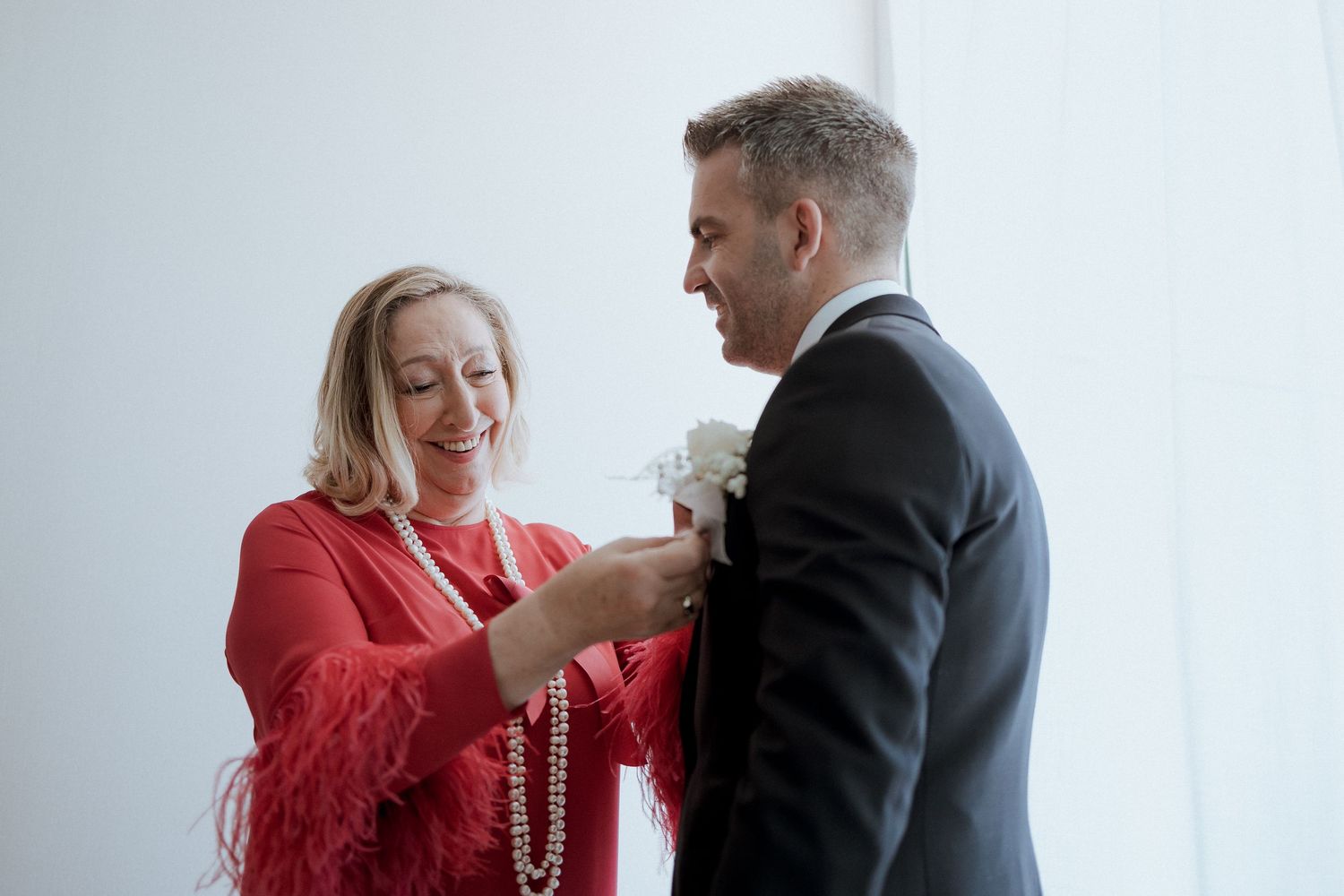 A smiling woman in a red feathered dress pins a white boutonniere on a man in a black suit.