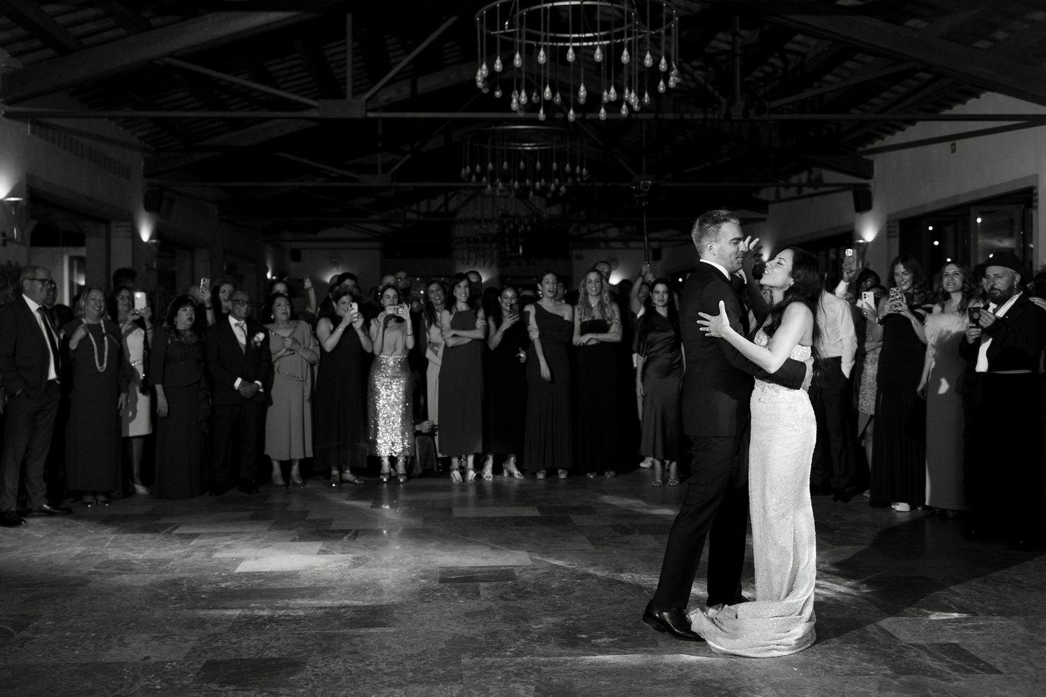 Bride and groom share first dance surrounded by guests in elegant black and white photo.