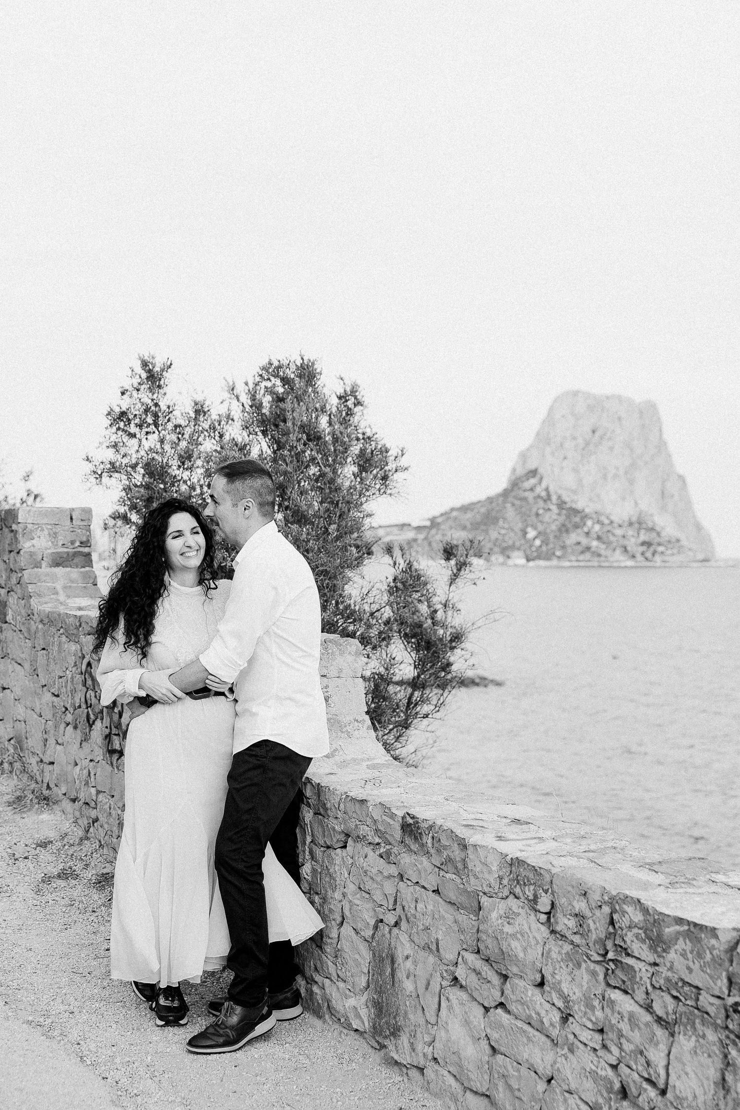 A couple shares a romantic moment on a stone wall overlooking a dramatic coastal landscape with a mountain in Spain.