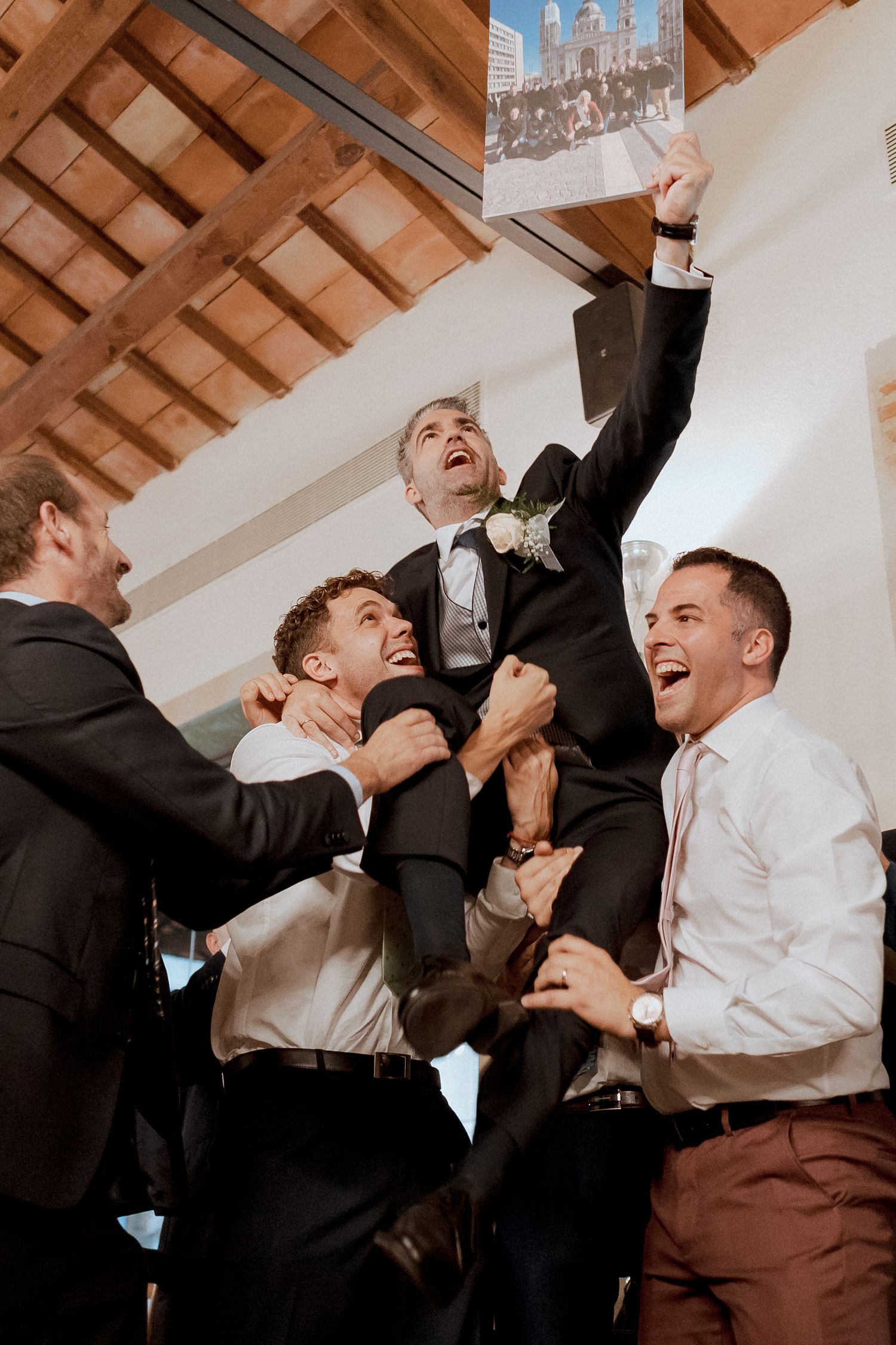 Groomsmen joyfully lifting the groom during a wedding celebration in a rustic venue.
