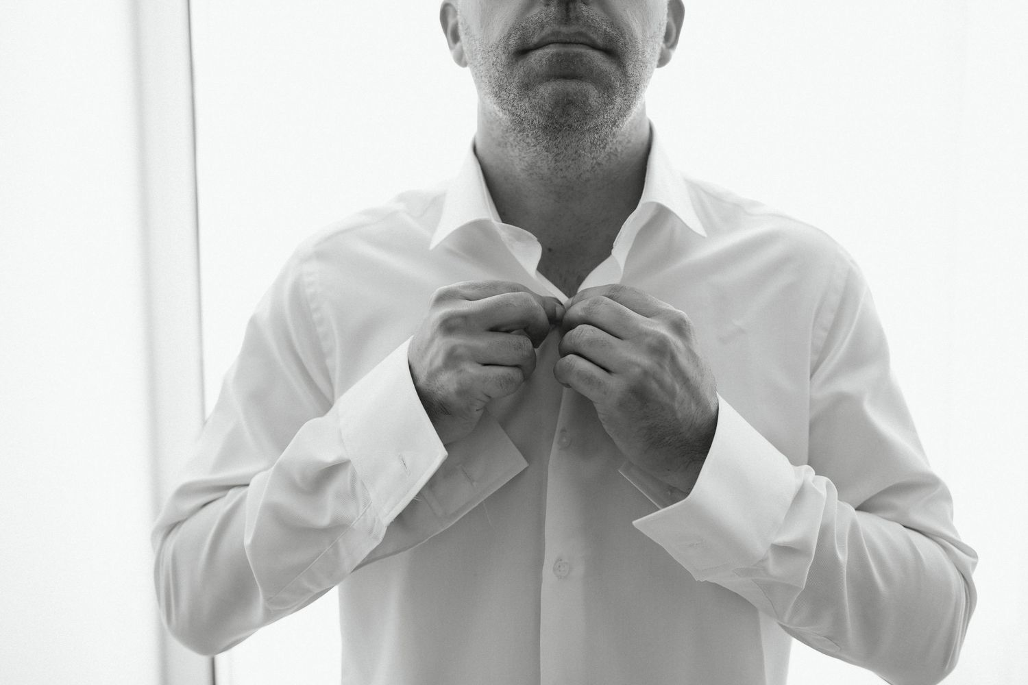 Black and white photo of a man in a white dress shirt buttoning his collar, getting dressed for a formal event.