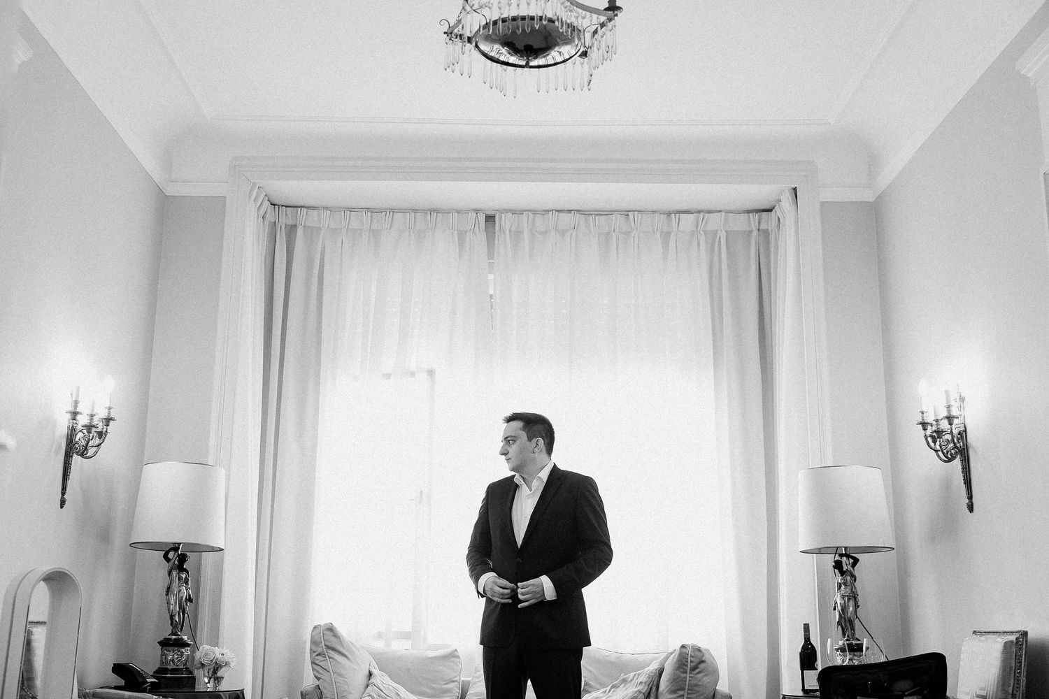 Groom in black suit standing by sheer curtains in a bright, classic room preparing for his wedding.