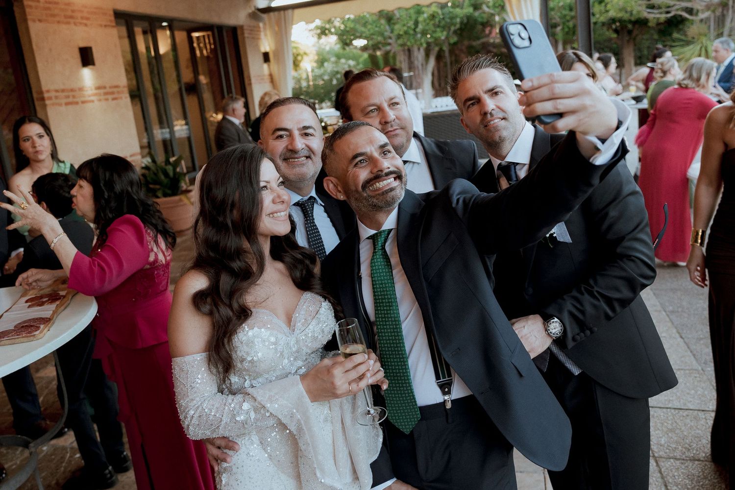 Wedding group selfie with bride in lace gown and groom in suit with green tie celebrating outdoors.
