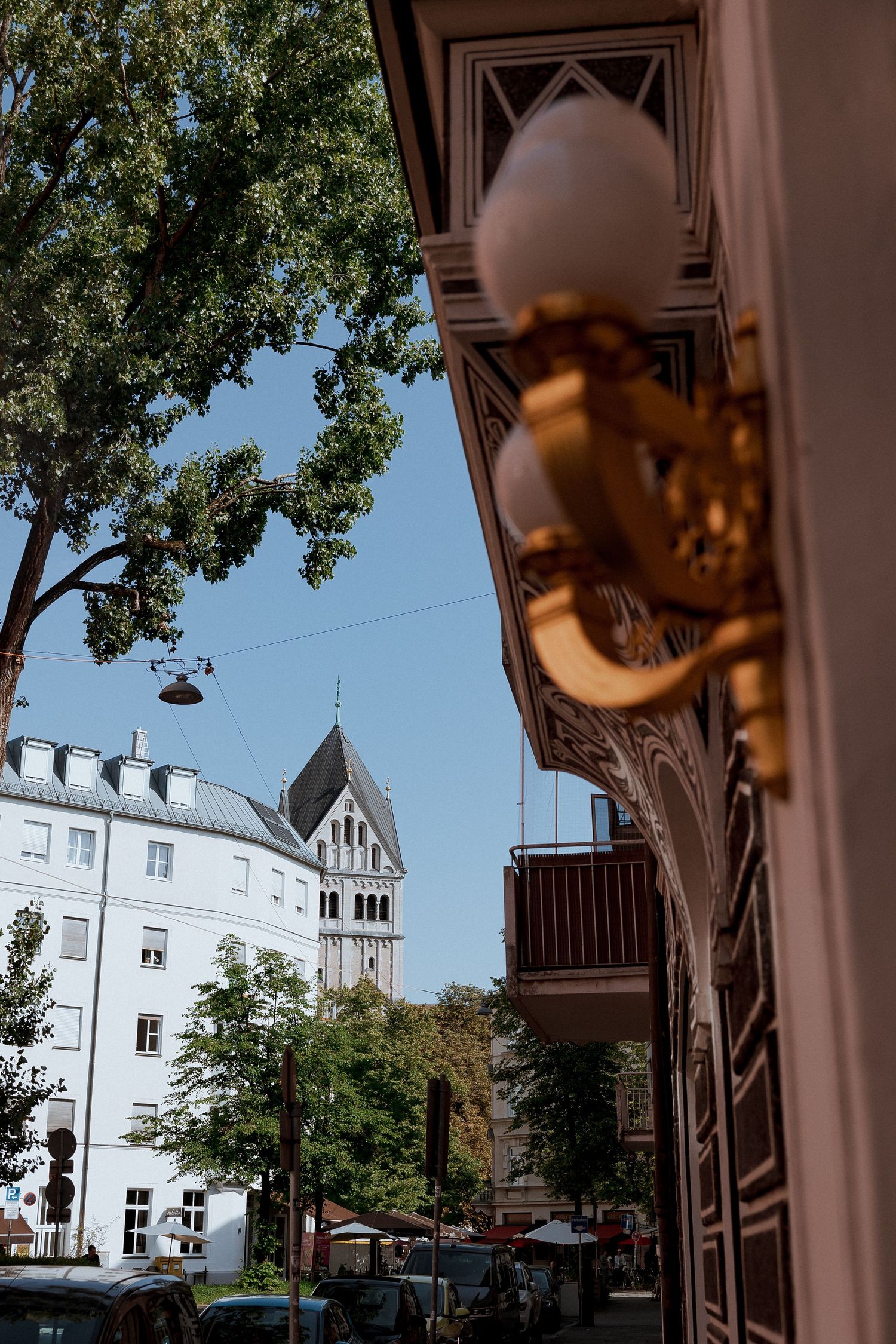 Golden wall sconce on ornate building facade with European street and historic church tower in background.