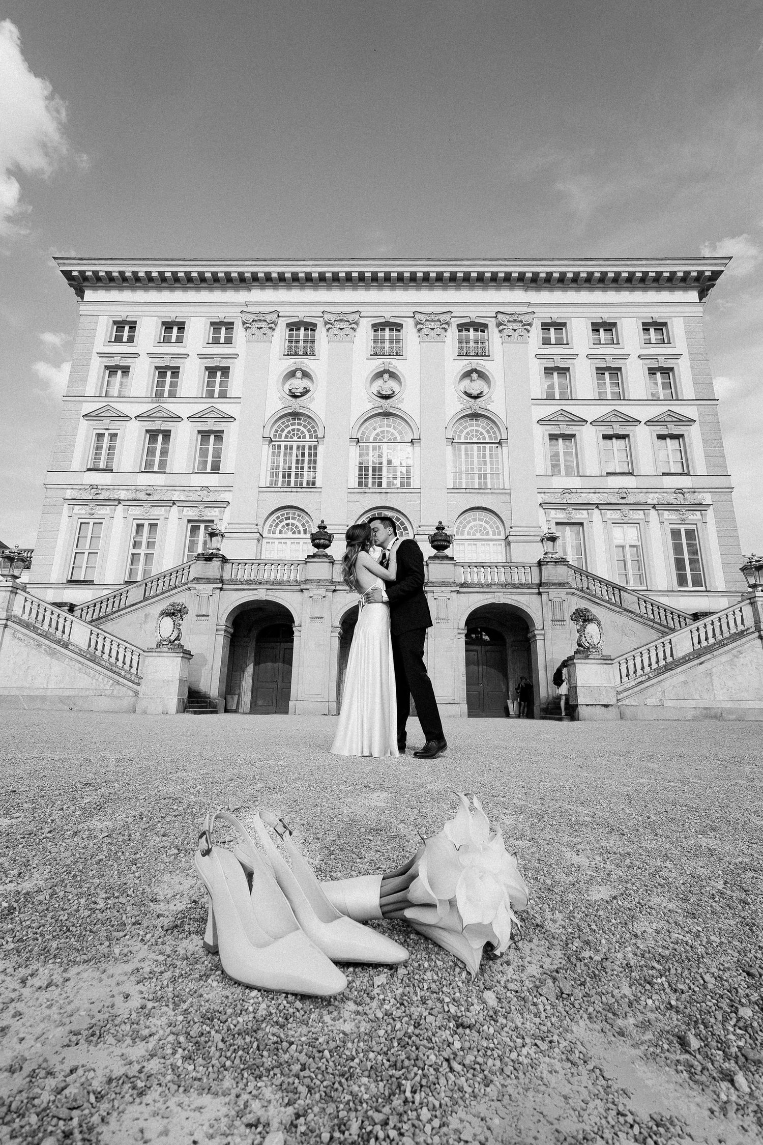 Black and white photo of newlyweds kissing before a grand neoclassical building with bridal shoes in foreground.