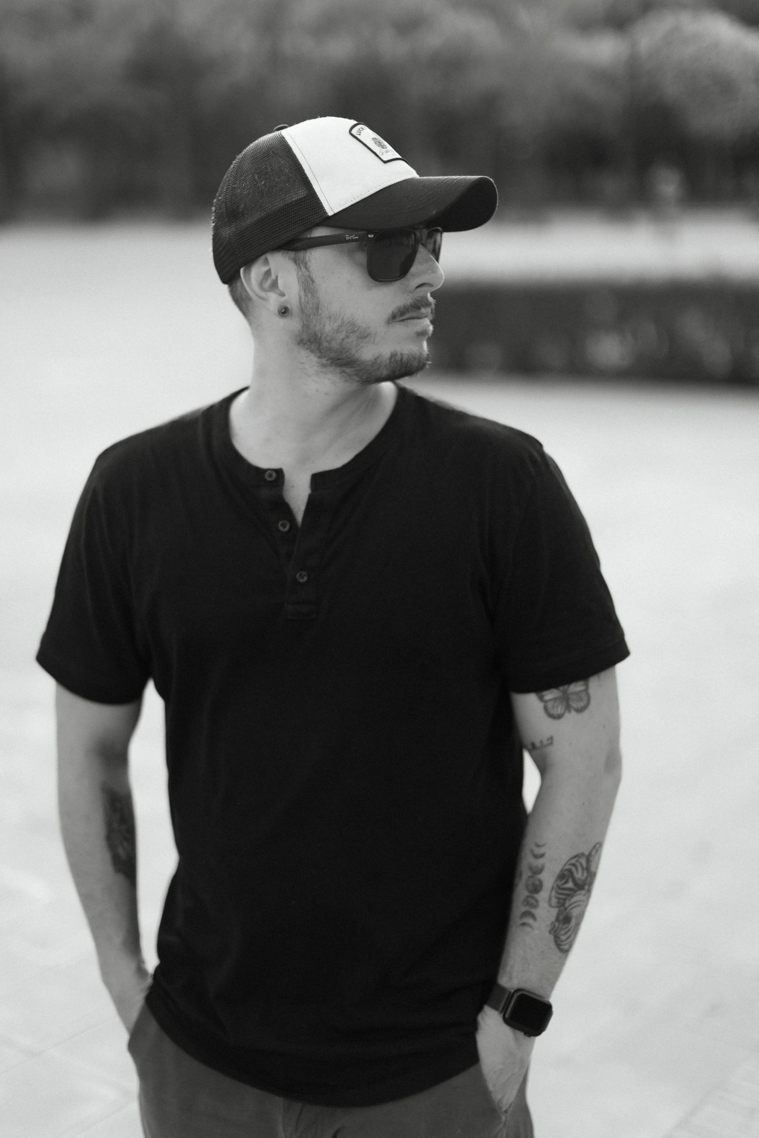 Black and white photo of a tattooed man in sunglasses, black Henley shirt, and trucker cap outdoors.