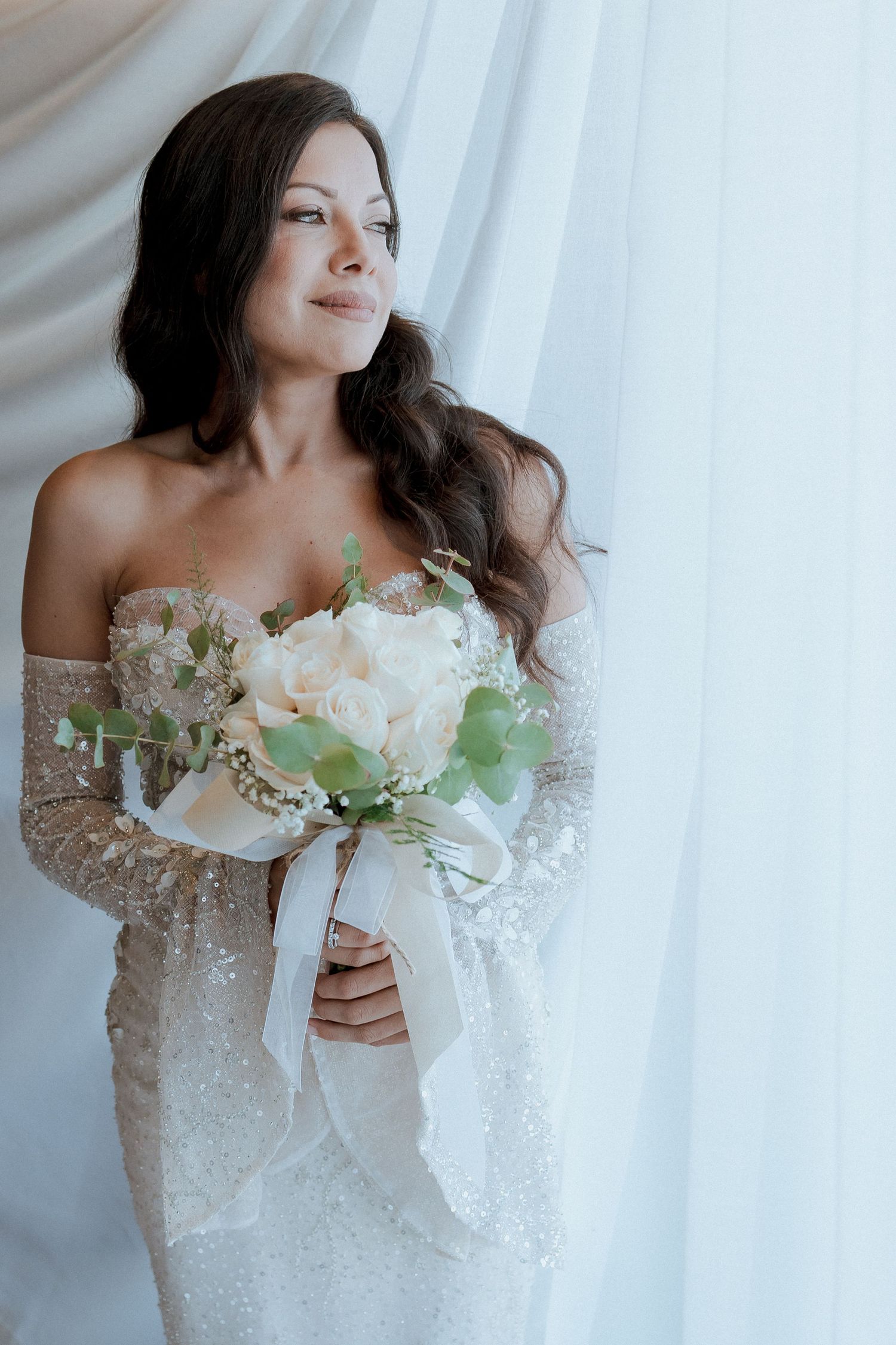 Elegant bride in a beaded off-shoulder gown holding a white rose bouquet, standing by sheer curtains in soft natural light.