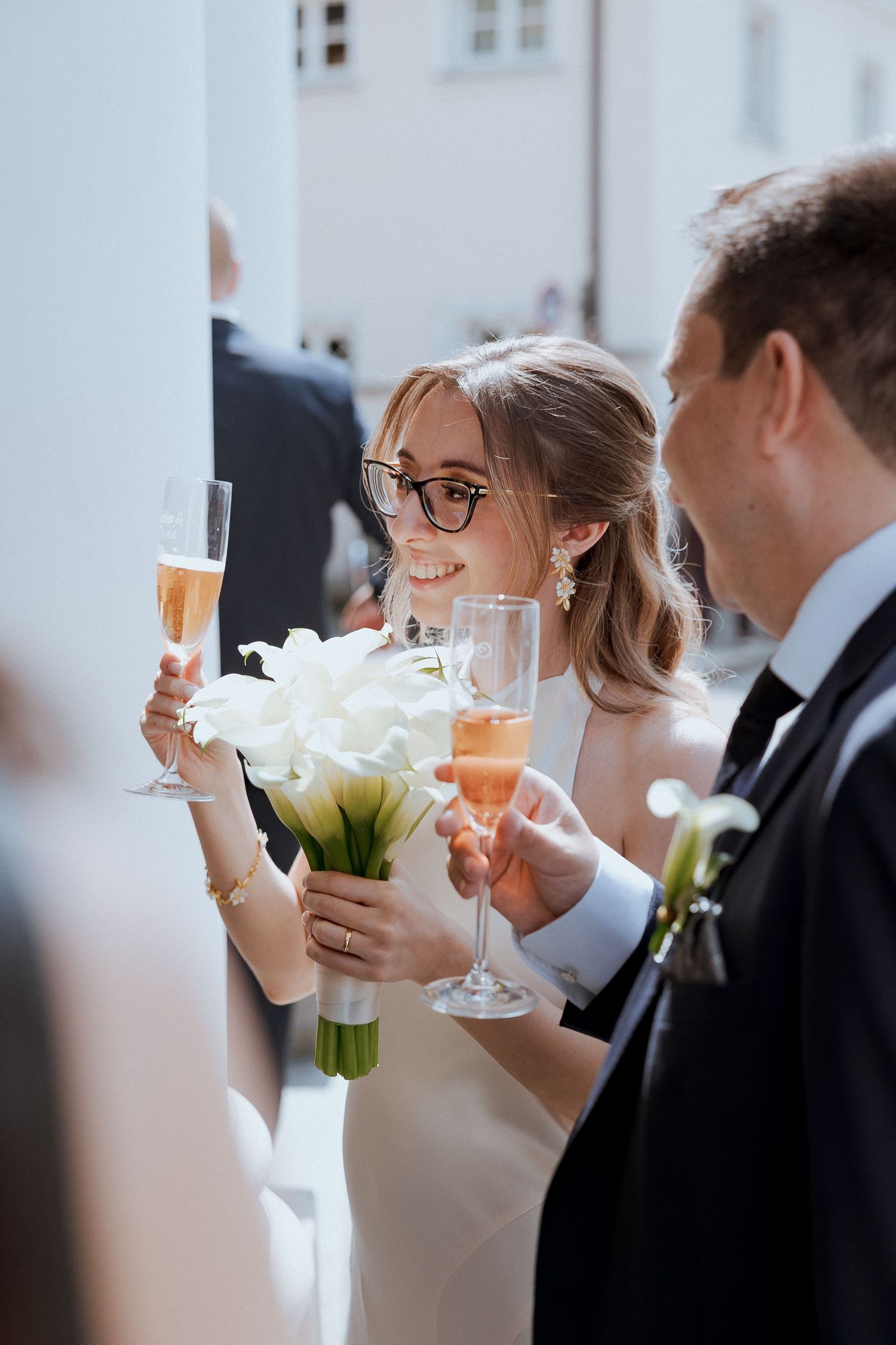 Smiling bride with glasses toasting champagne with groom while holding white calla lily bouquet.
