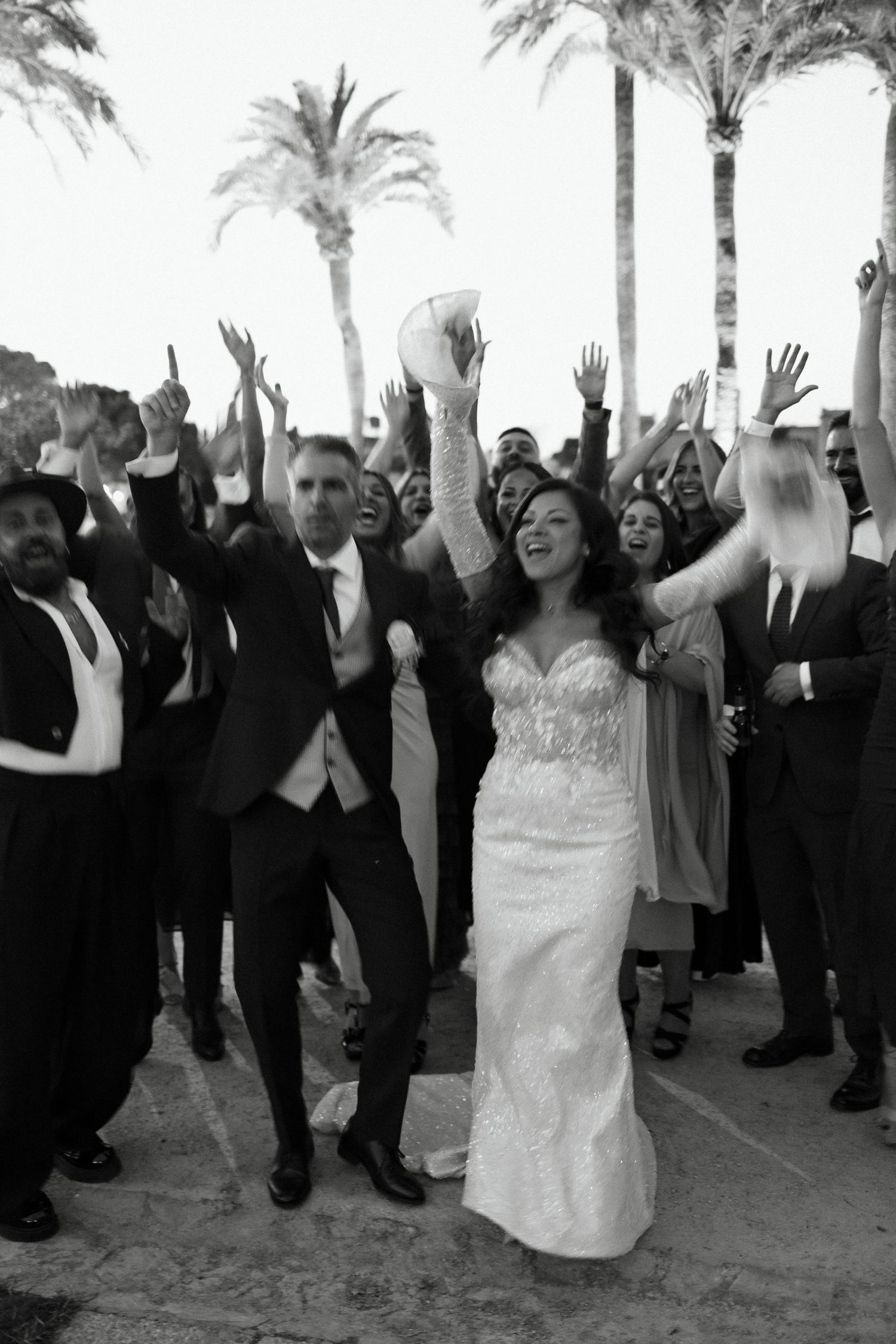 Bride and groom celebrate joyfully with wedding guests under palm trees in black and white photo.