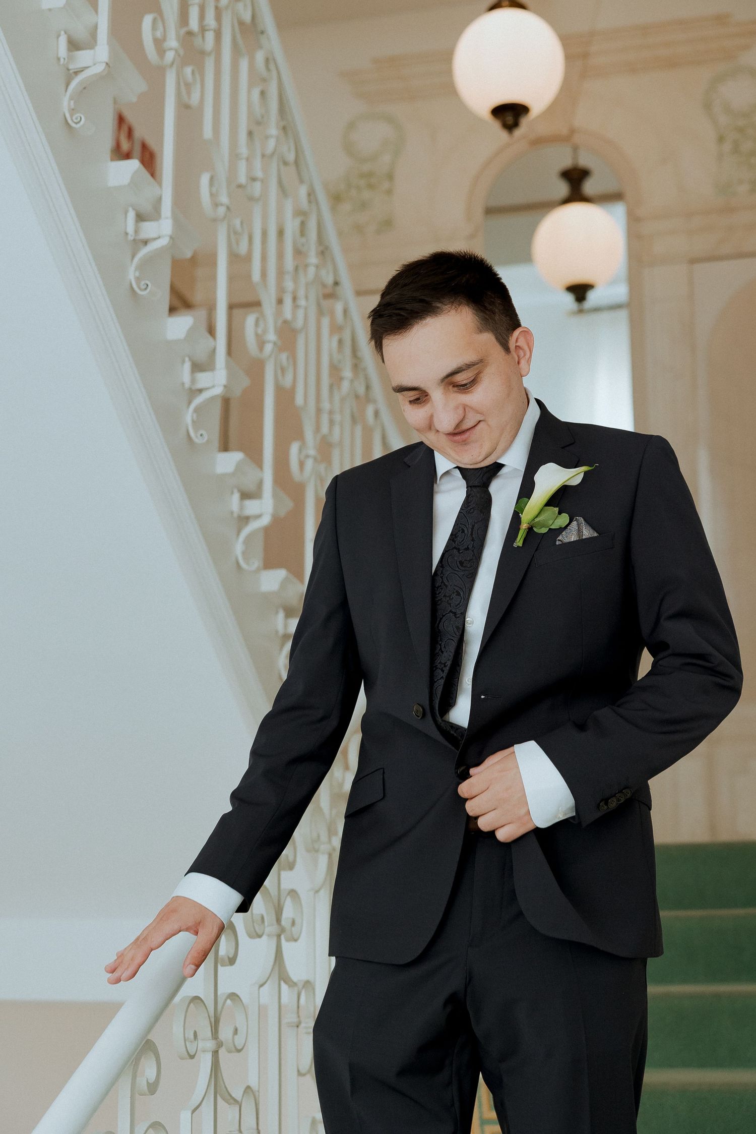 Elegant groom in black suit with white boutonniere posing on ornate staircase in classic venue.