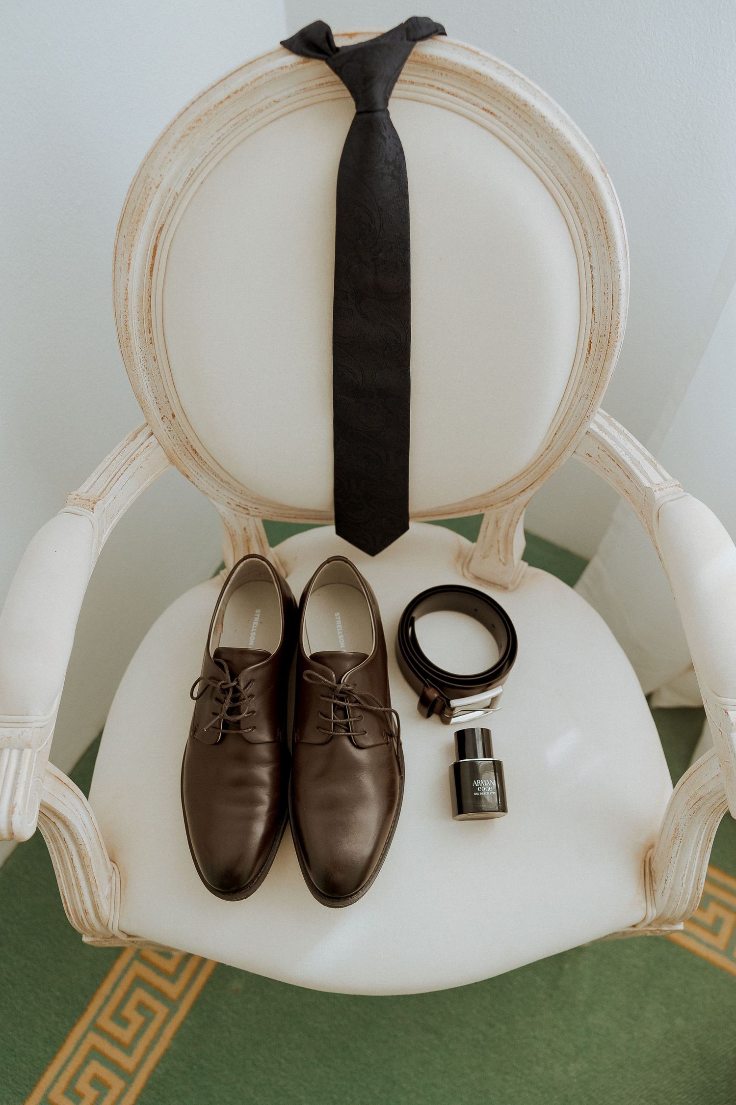 Groom wedding accessories flat lay with brown oxford shoes, black tie, belt, and cologne on white chair.