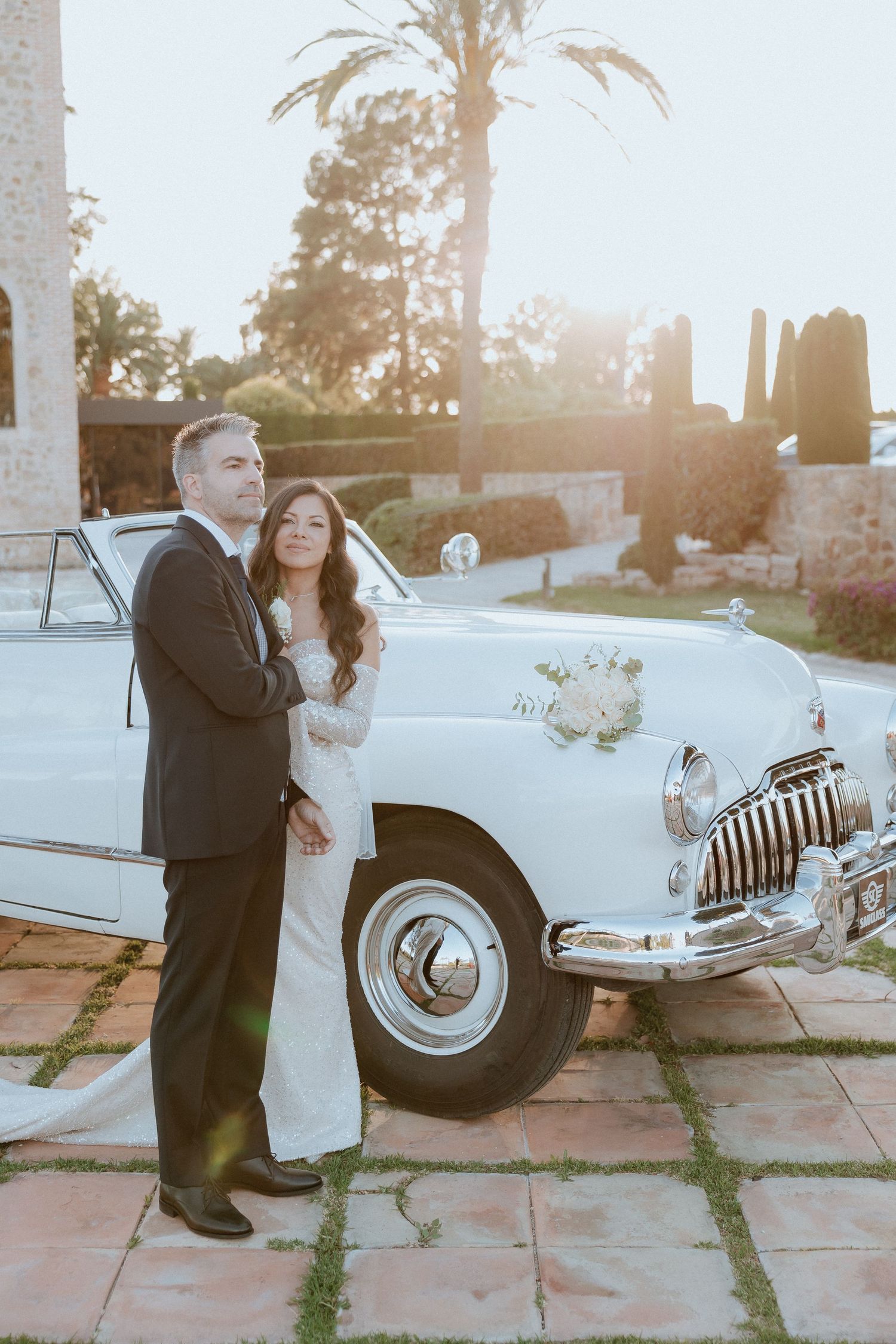 Bride and groom posing beside a vintage white Buick classic car at golden hour, surrounded by palm trees.