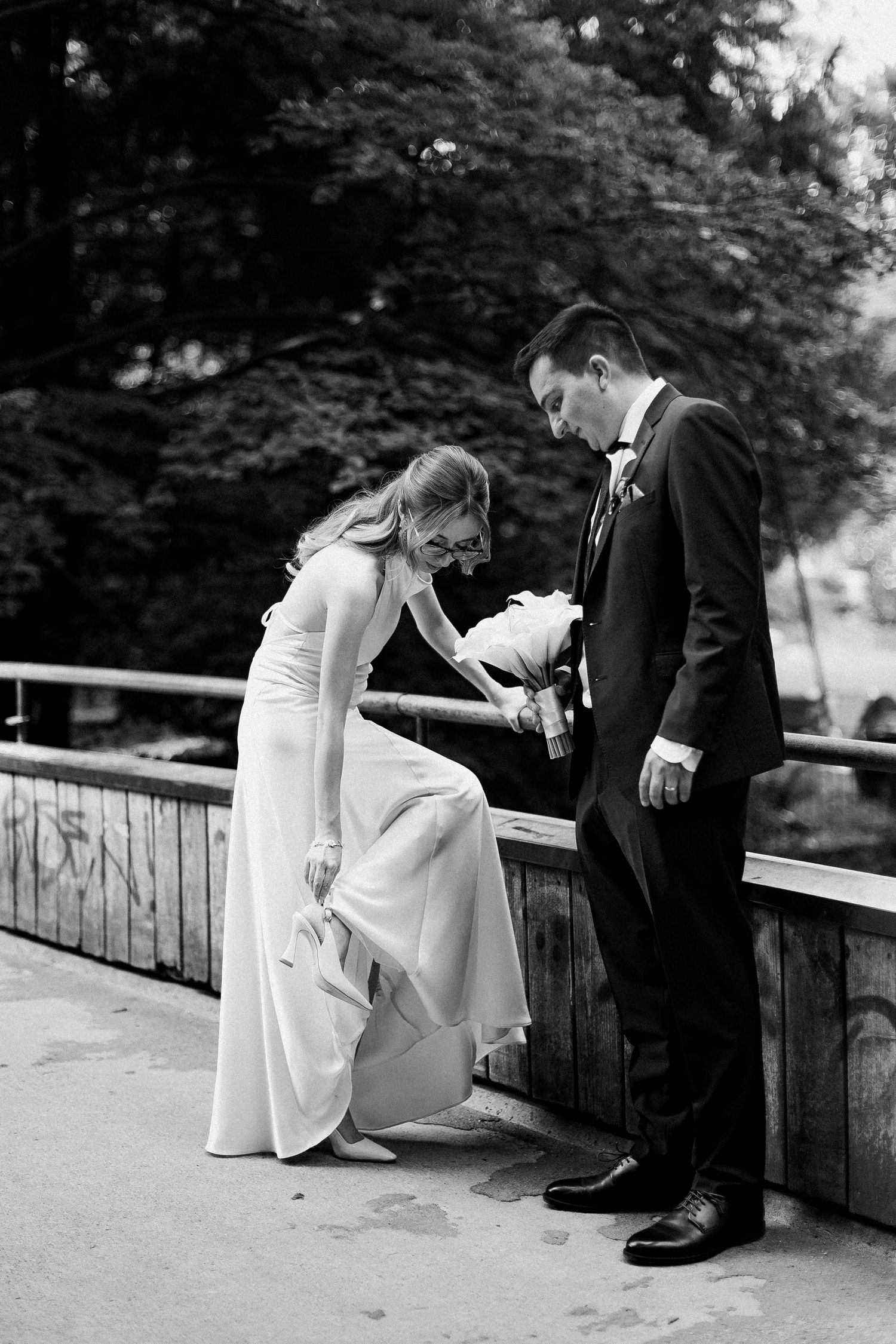 Bride in white gown and groom in black tuxedo share a tender moment on a bridge in black and white photo.
