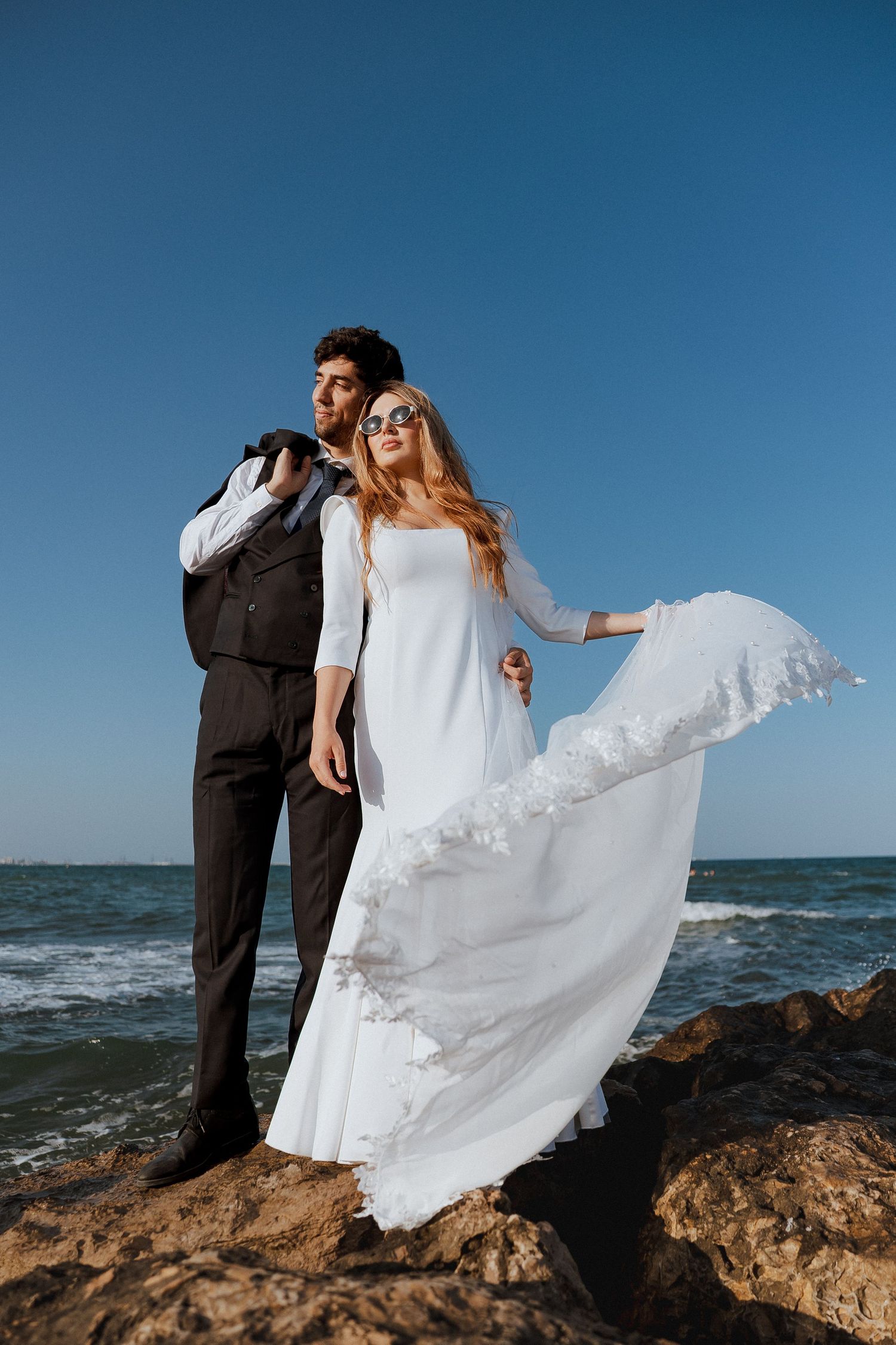 A couple stands on coastal rocks as the bride's white dress flows dramatically in the ocean breeze.