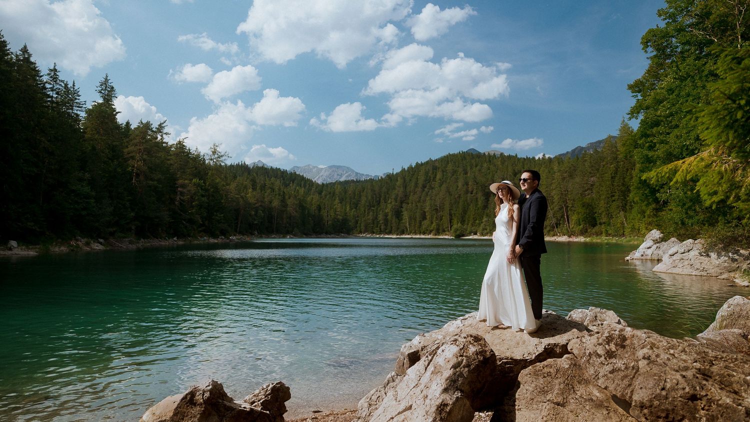 A panoramic mountain landscape with turquoise lake waters surrounded by evergreen forests under blue skies.