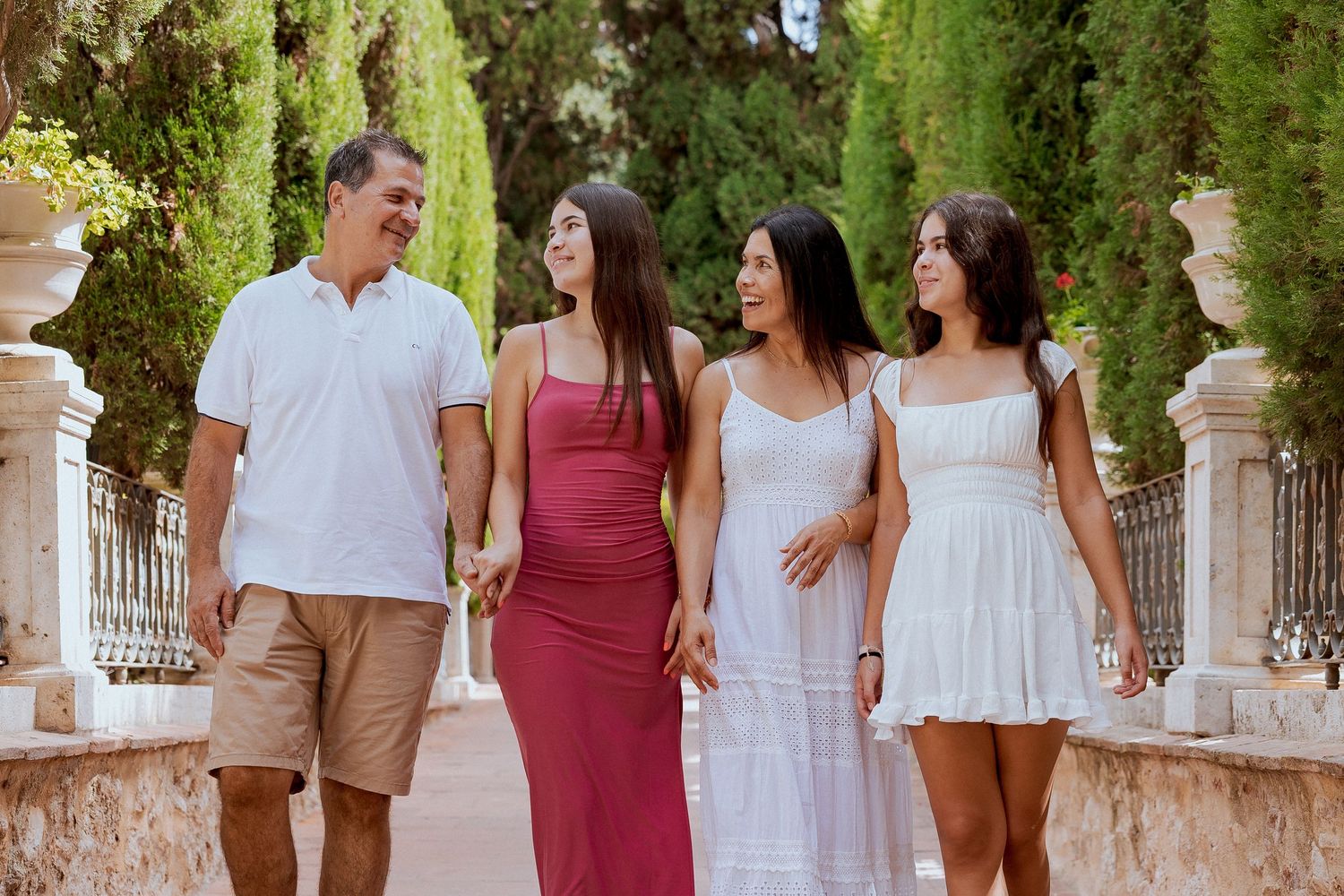 Wedding party walking together through a garden path lined with cypress trees in white and burgundy attire.