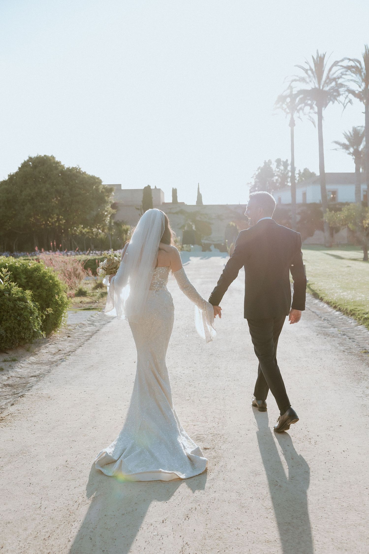 Bride and groom walk hand in hand down sunlit garden path, bride wearing flowing veil and fitted white gown.