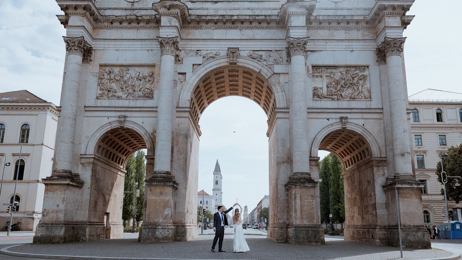Bride and groom dancing under a grand ornate stone arch monument in a European city square.
