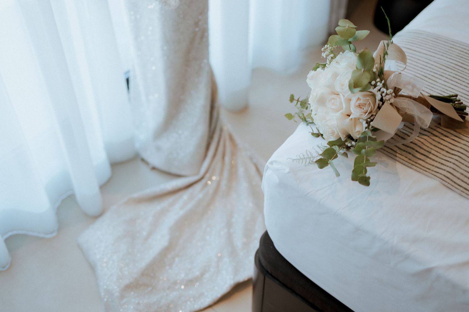 White lace wedding dress draped beside a floral bouquet of roses resting on a striped surface.