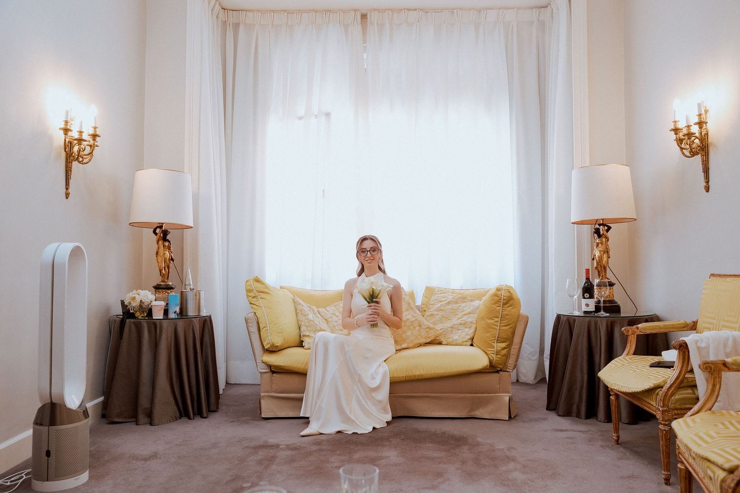 Bride in white wedding dress sitting on yellow sofa in elegant hotel suite with gold wall sconces.