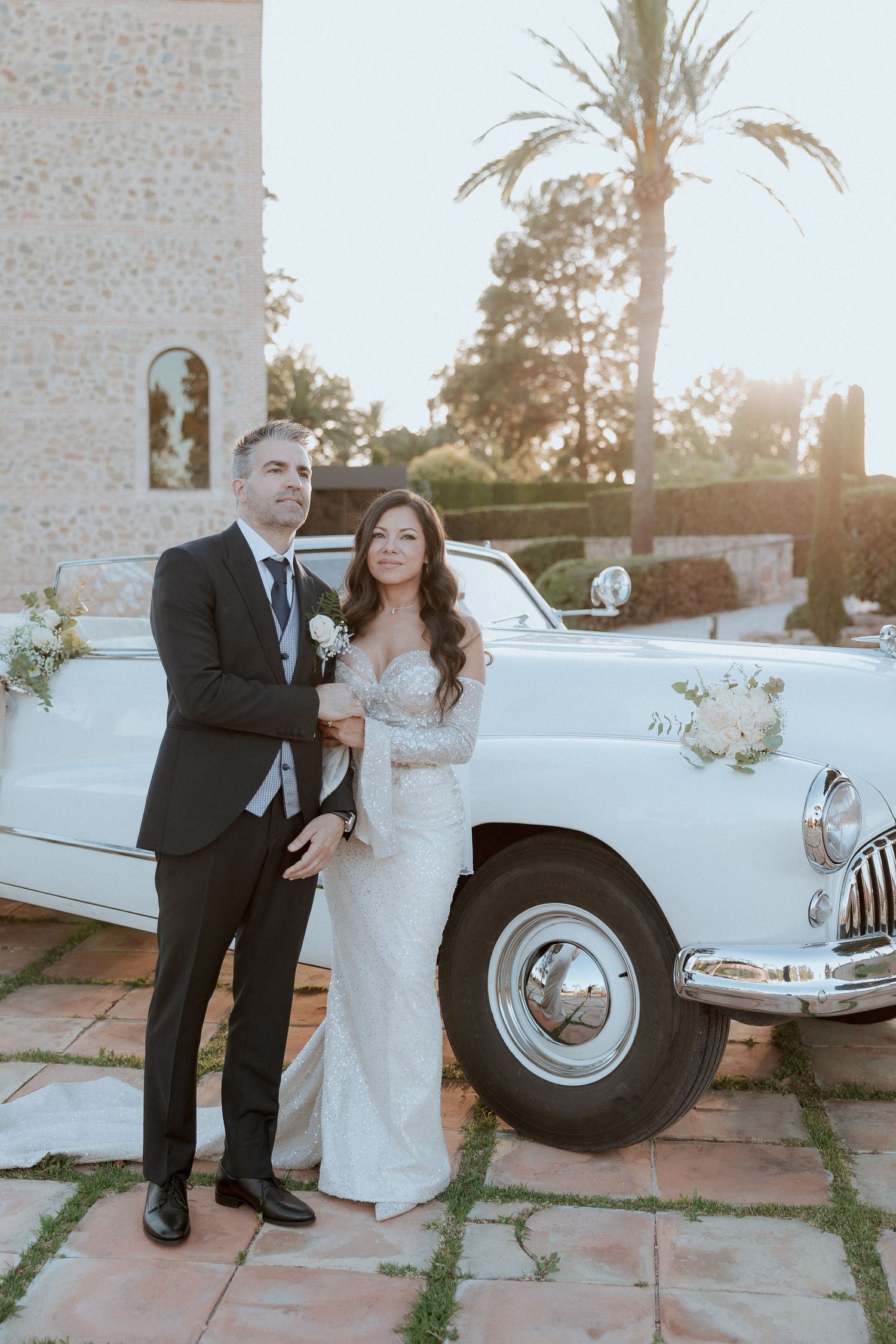 Bride and groom pose beside a classic white vintage car outdoors with palm trees and golden sunlight behind them.
