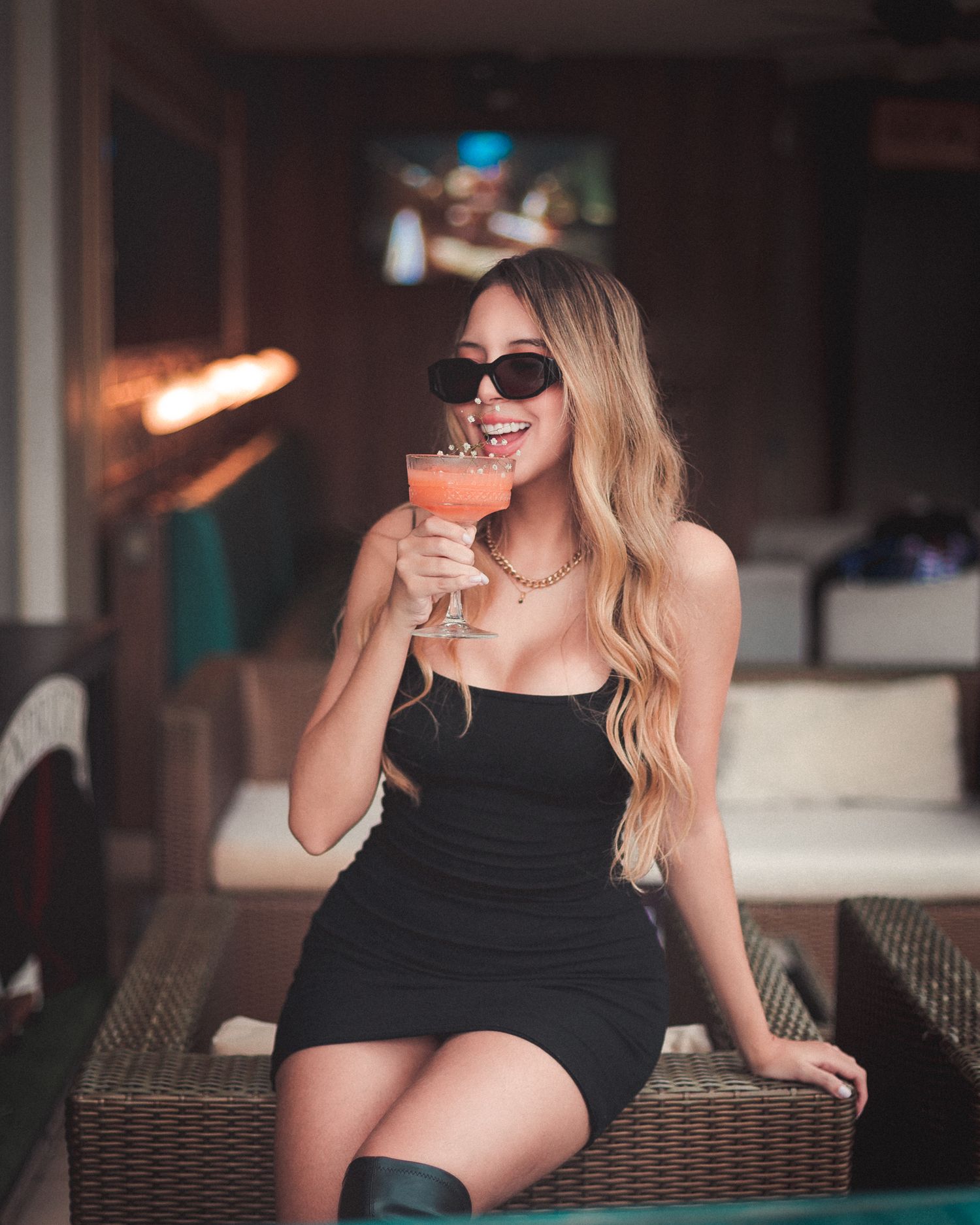 Woman in black dress sips cocktail at bar counter while relaxing in elegant evening setting.