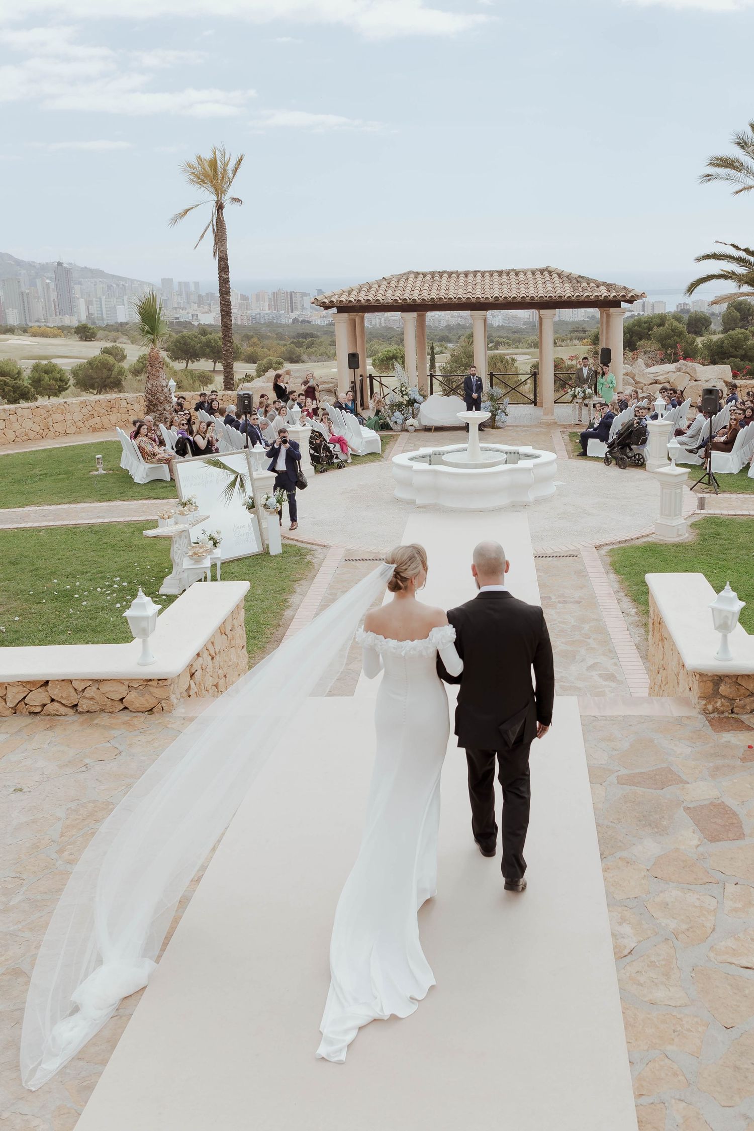 Bride in white gown with long veil walks down outdoor aisle with escort toward gazebo ceremony venue.
