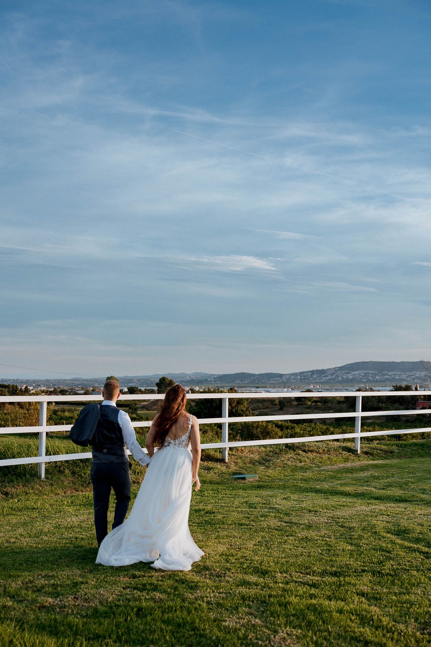 Bride and groom holding hands walking along white fence overlooking scenic countryside landscape on their wedding day.