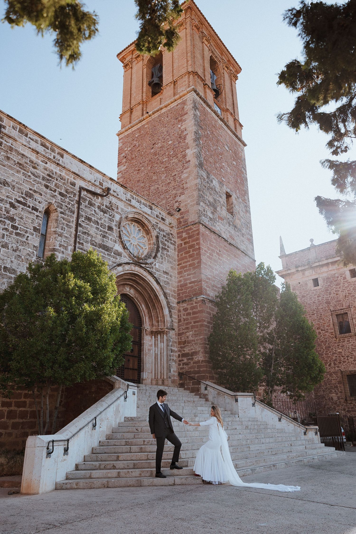A couple poses on stone steps leading to a historic church with a tall brick bell tower at sunset.