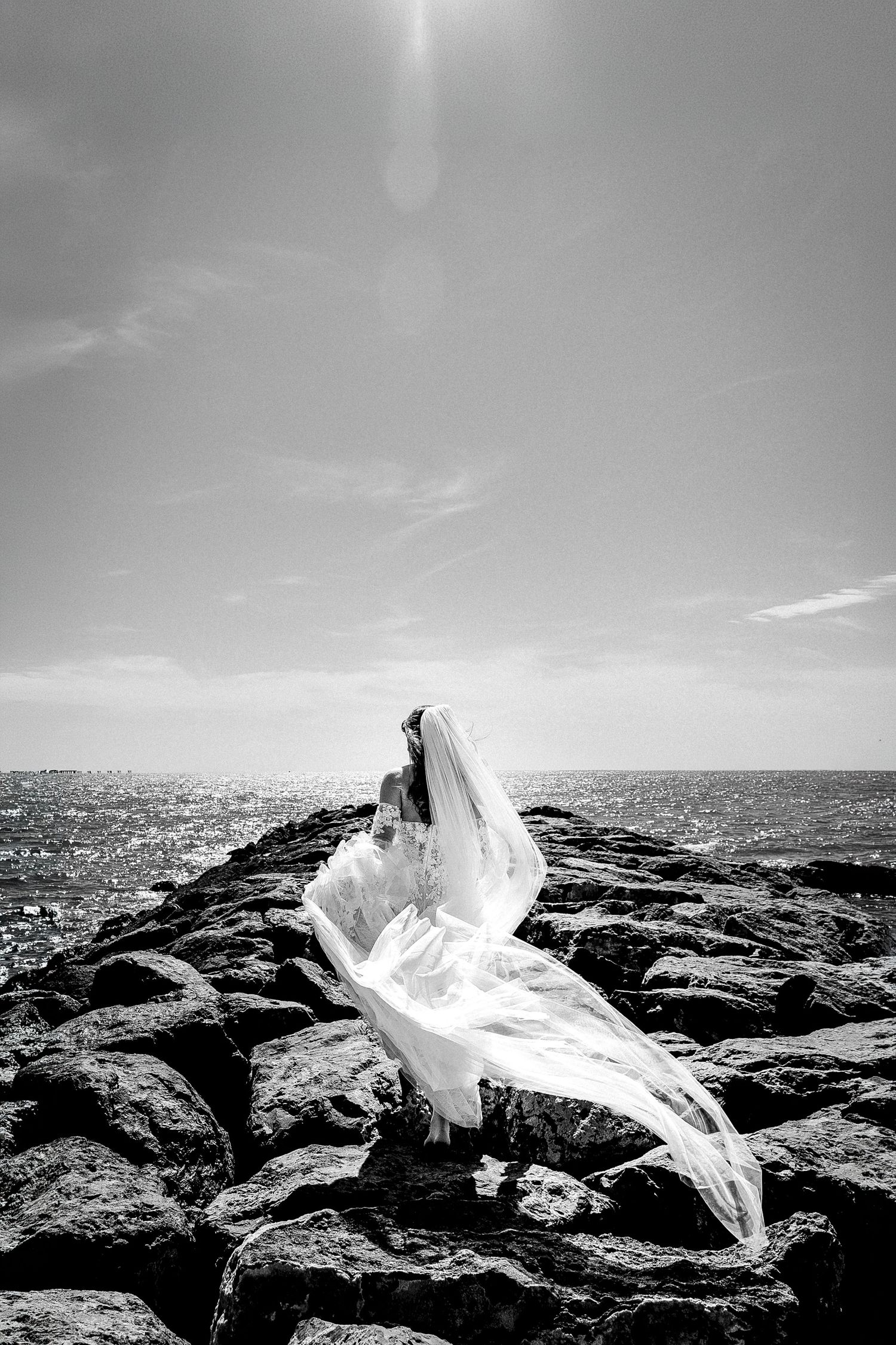 Black and white photo of a bride in flowing wedding dress and veil standing on rocky shoreline with ocean backdrop.
