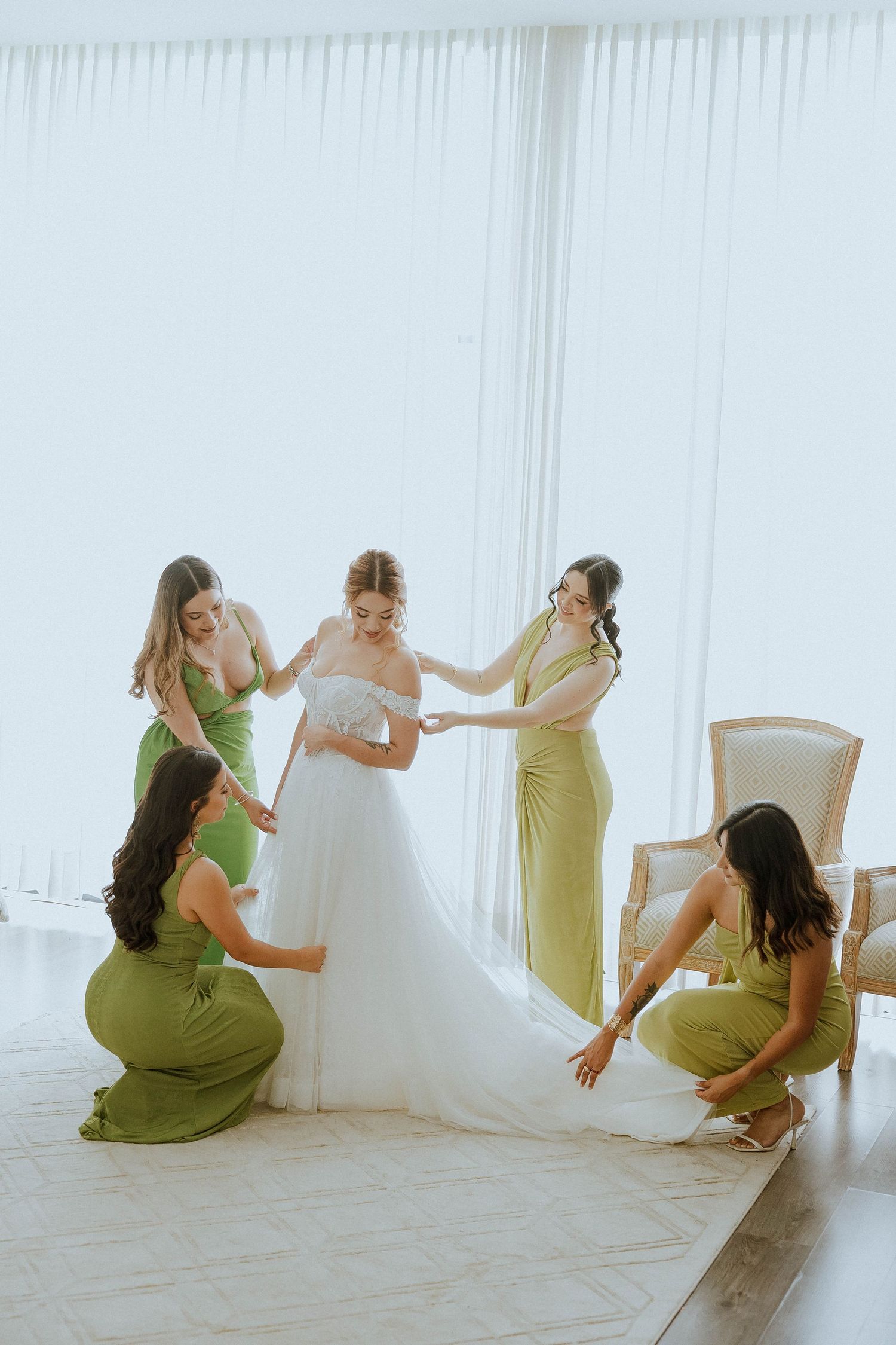 Bridesmaids in sage green dresses help adjust a white wedding gown in a bright studio setting.