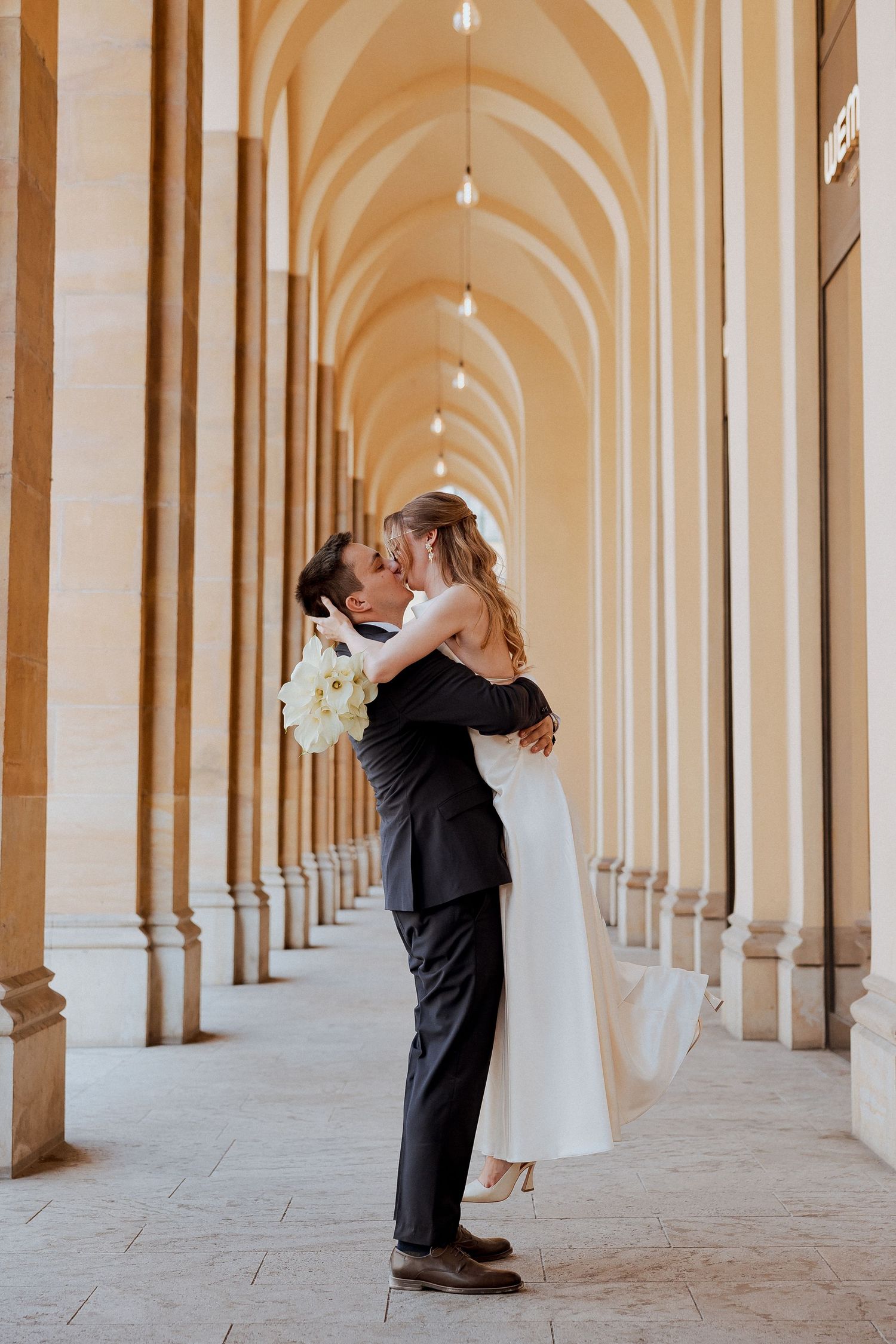 Bride and groom sharing a romantic kiss in an elegant arched corridor, bride holding white bouquet.