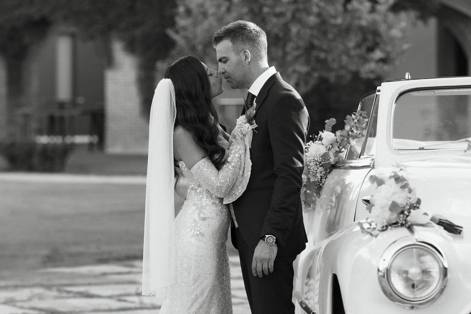 Black and white wedding photo of bride and groom sharing a romantic moment beside a vintage decorated car.
