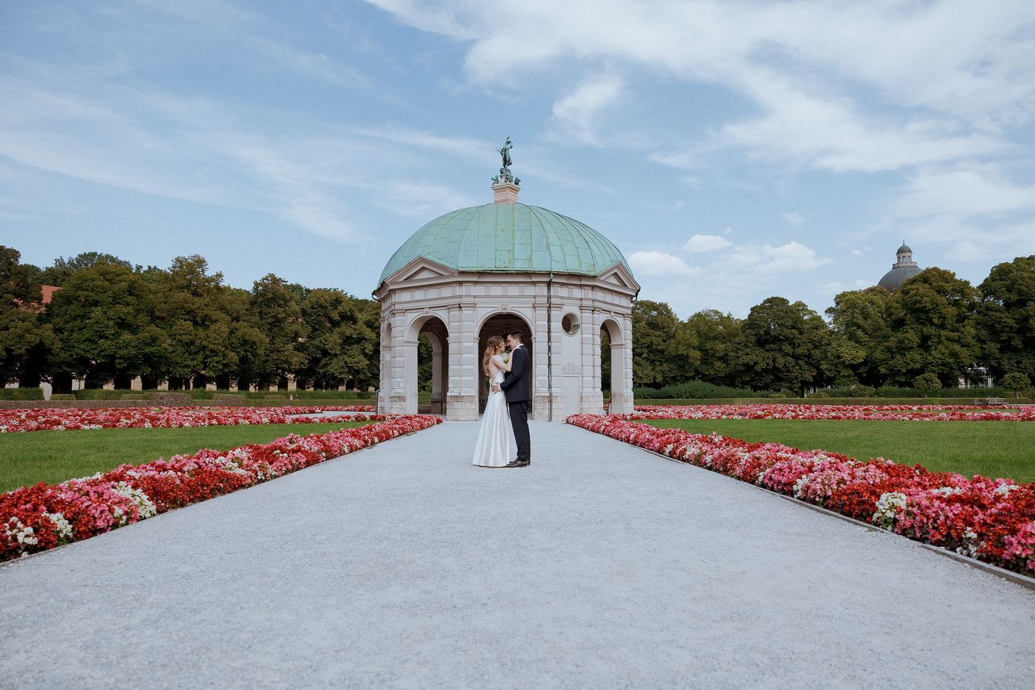 Wedding couple kissing at the Dianatempel in Munich's Hofgarten surrounded by vibrant red flower beds.