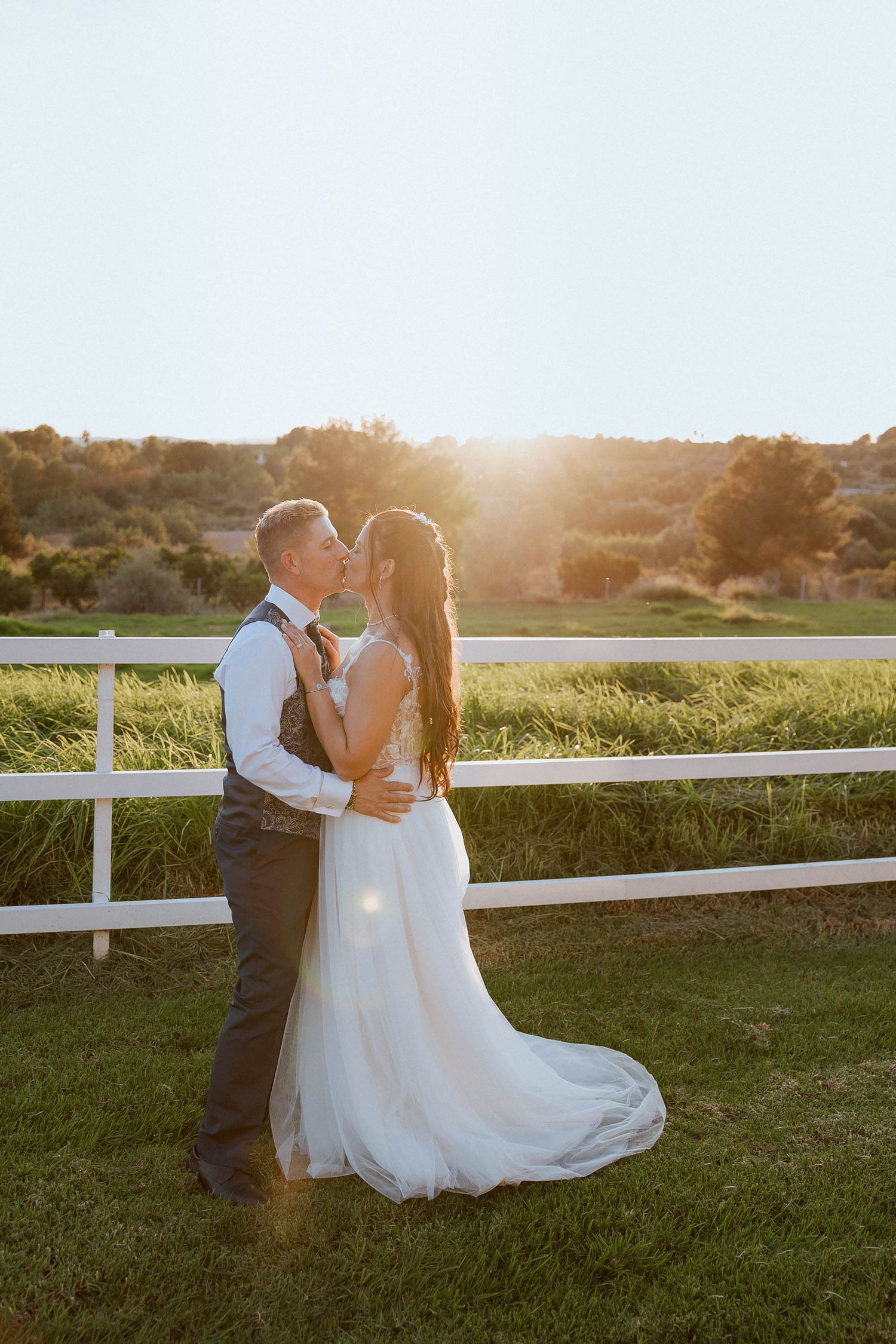 Newlywed couple shares romantic sunset moment by white fence at rustic countryside venue.
