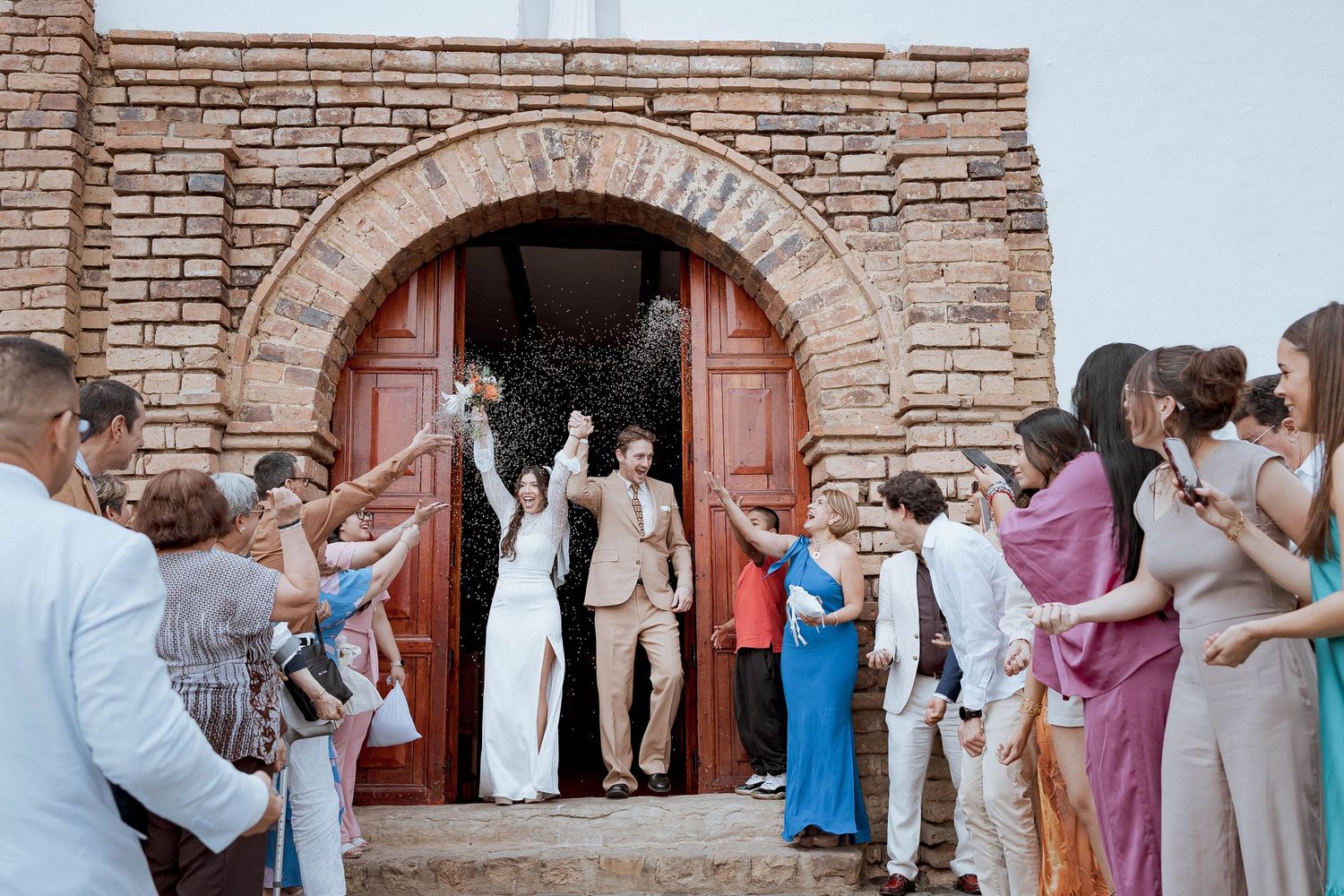 Wedding guests dance in a line formation against a rustic brick wall backdrop at an outdoor reception celebration.
