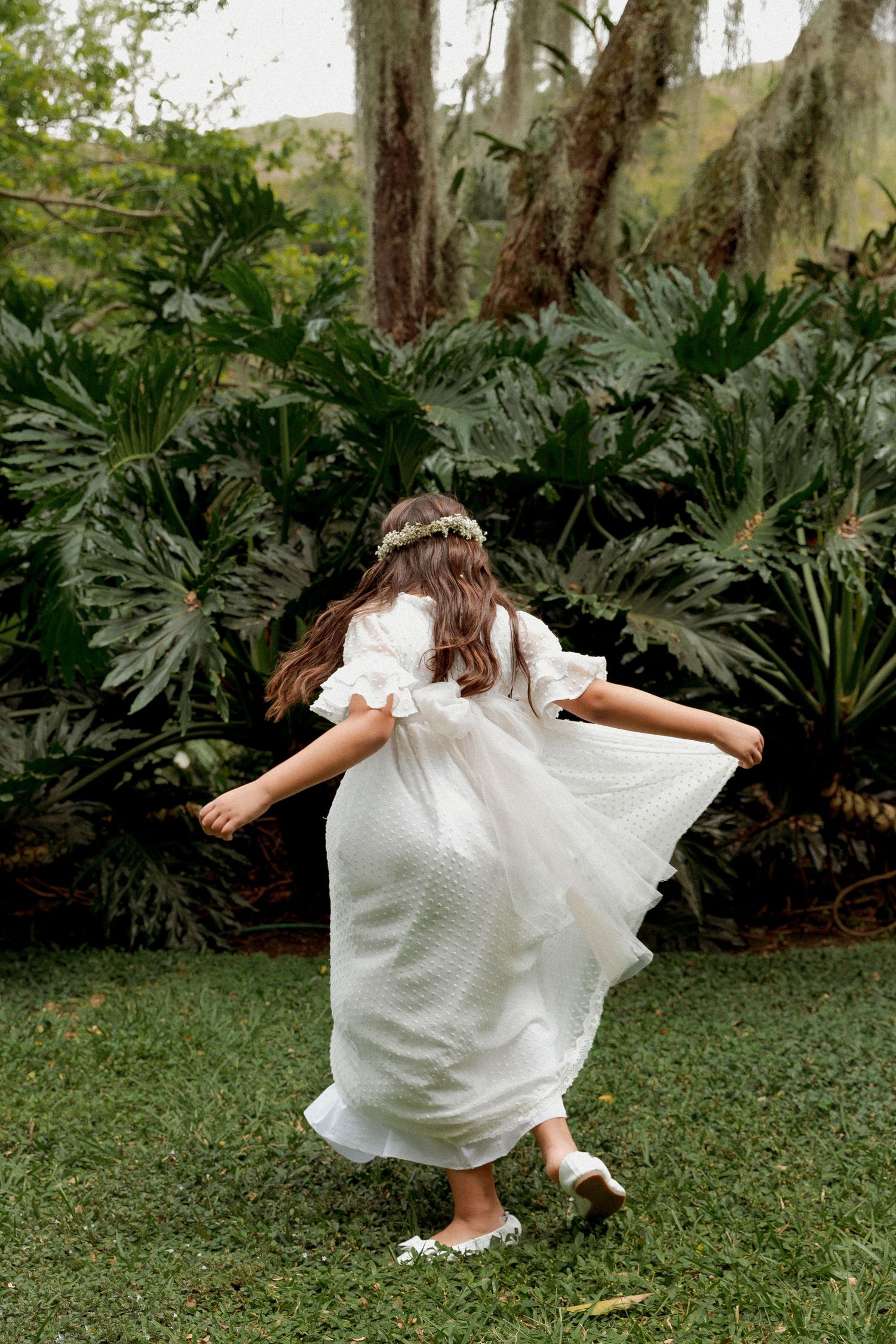 A person in a flowing white dress twirls playfully on a lush green lawn surrounded by tropical palm fronds and trees.