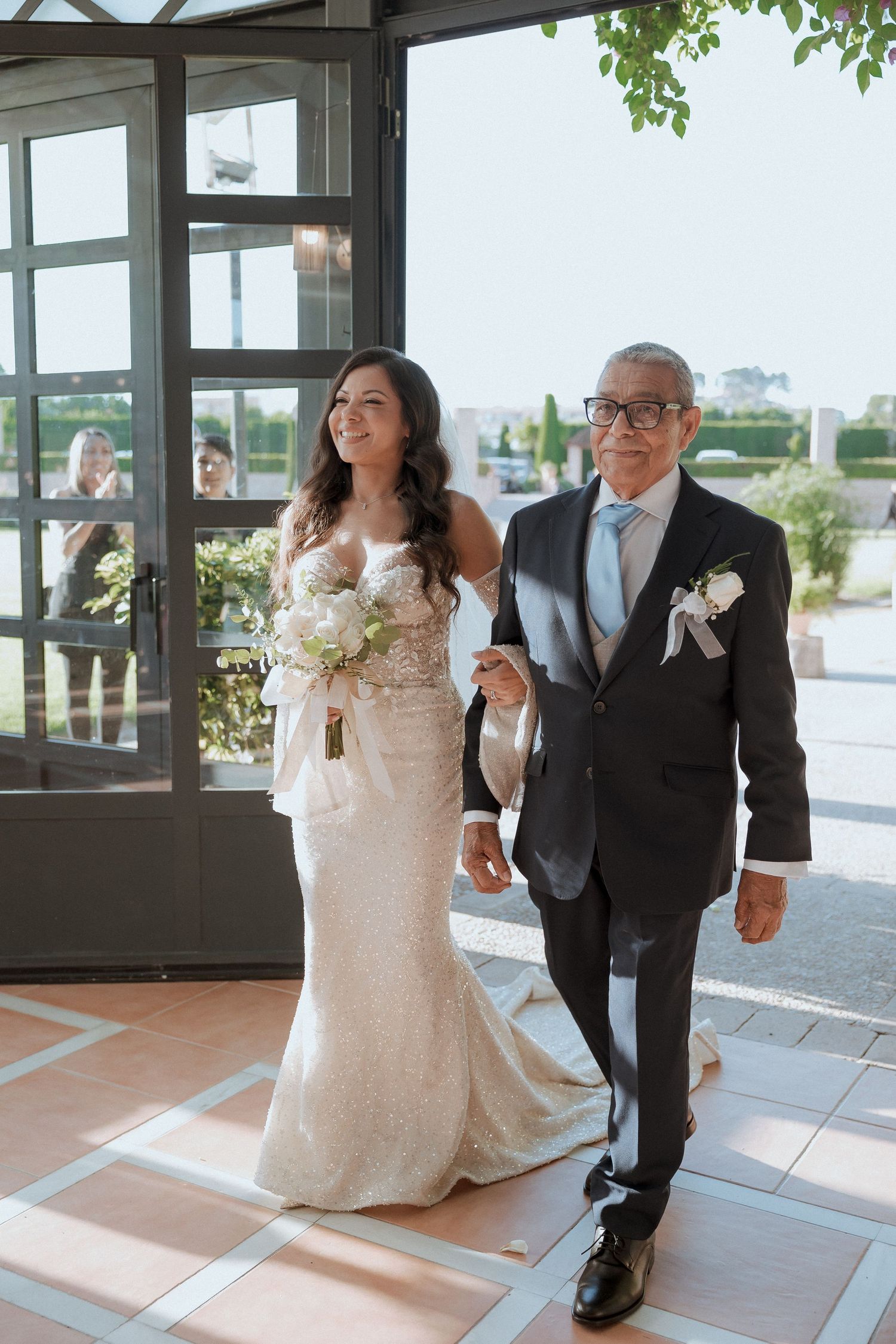 Bride in lace gown walks down aisle with older gentleman in dark suit and boutonniere at elegant wedding venue.