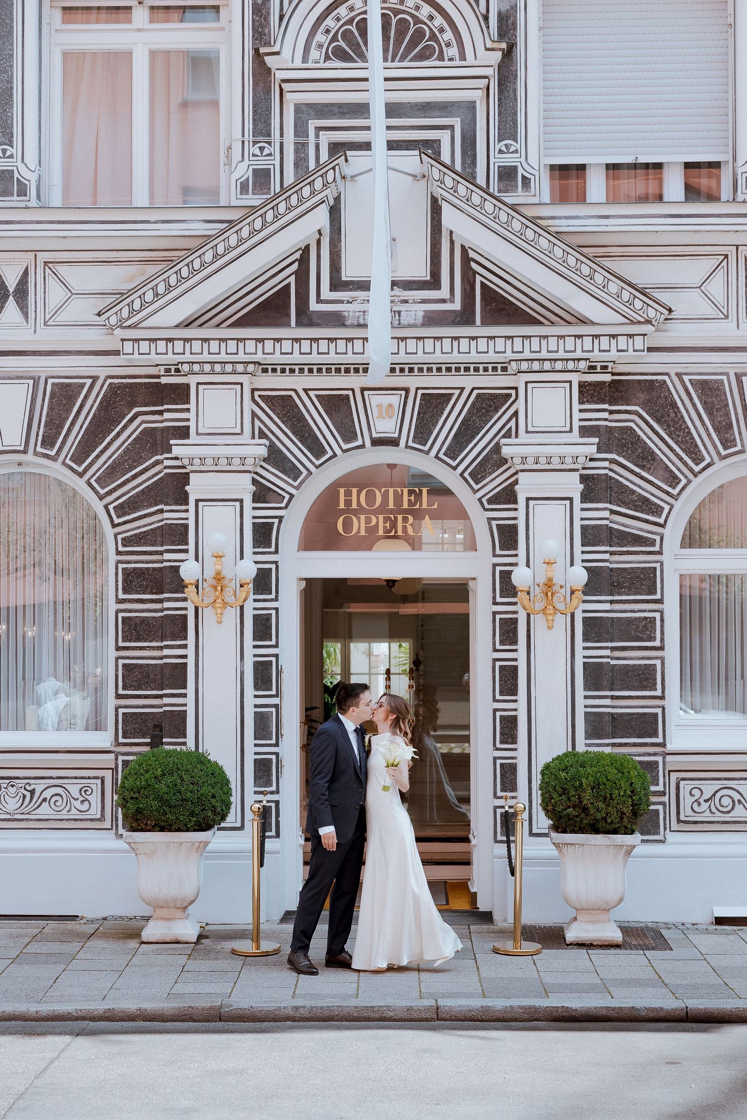 Wedding couple kissing in front of Hotel Oper facade in classic European architectural setting.