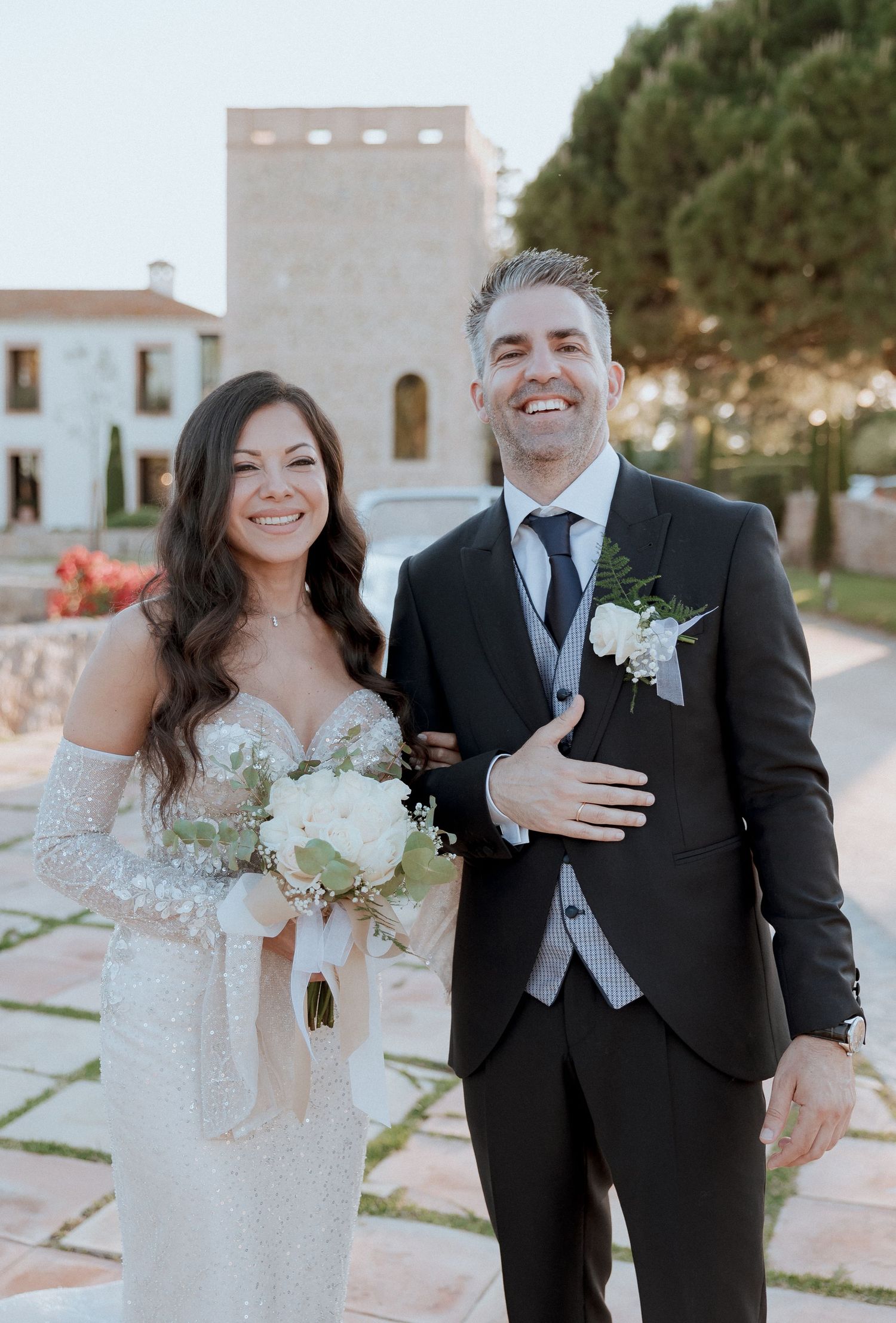 Smiling bride and groom pose outdoors at stone wedding venue, groom in black suit with white boutonniere.