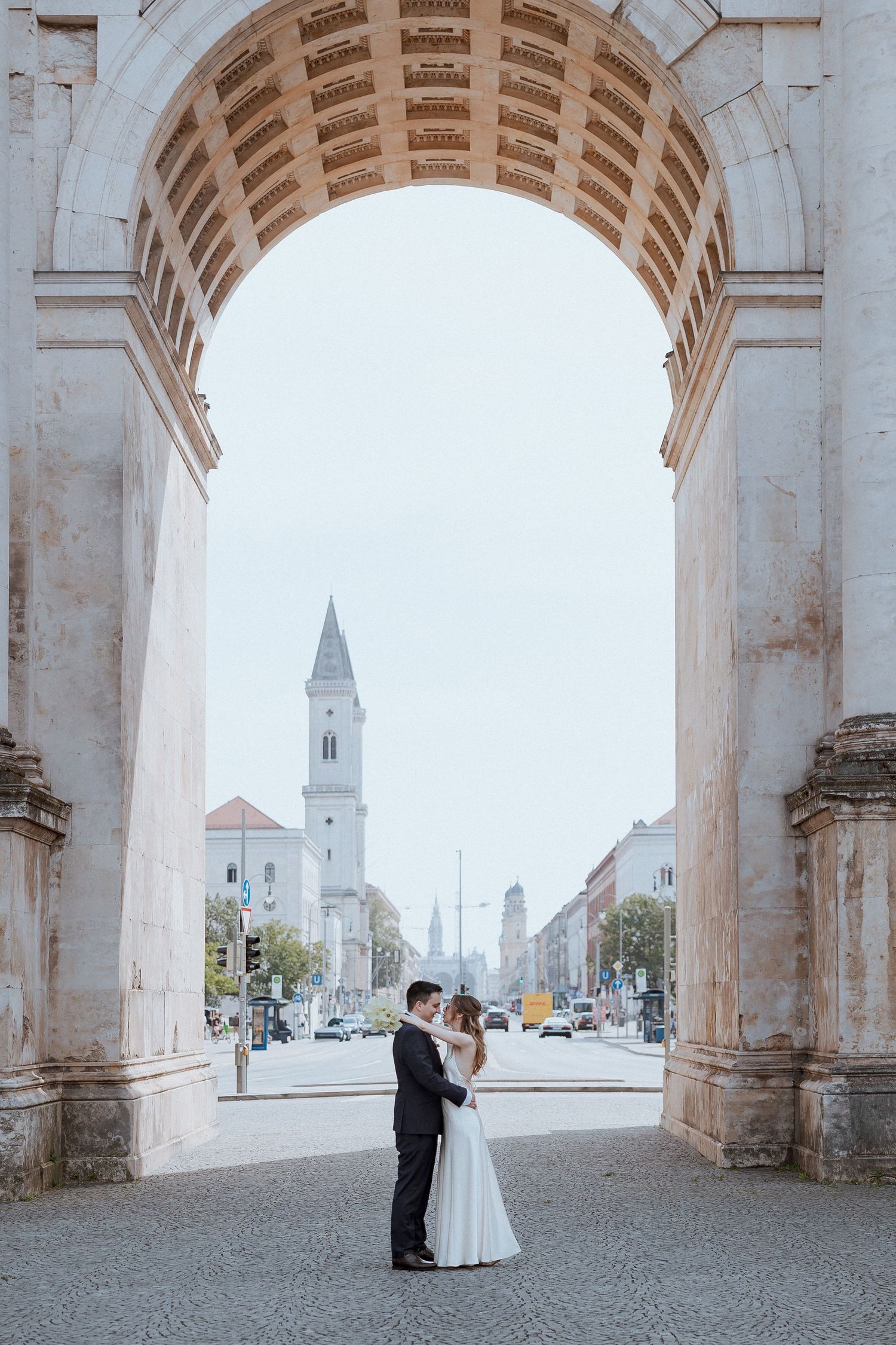 Wedding couple embracing under historic stone arch with church tower in background.