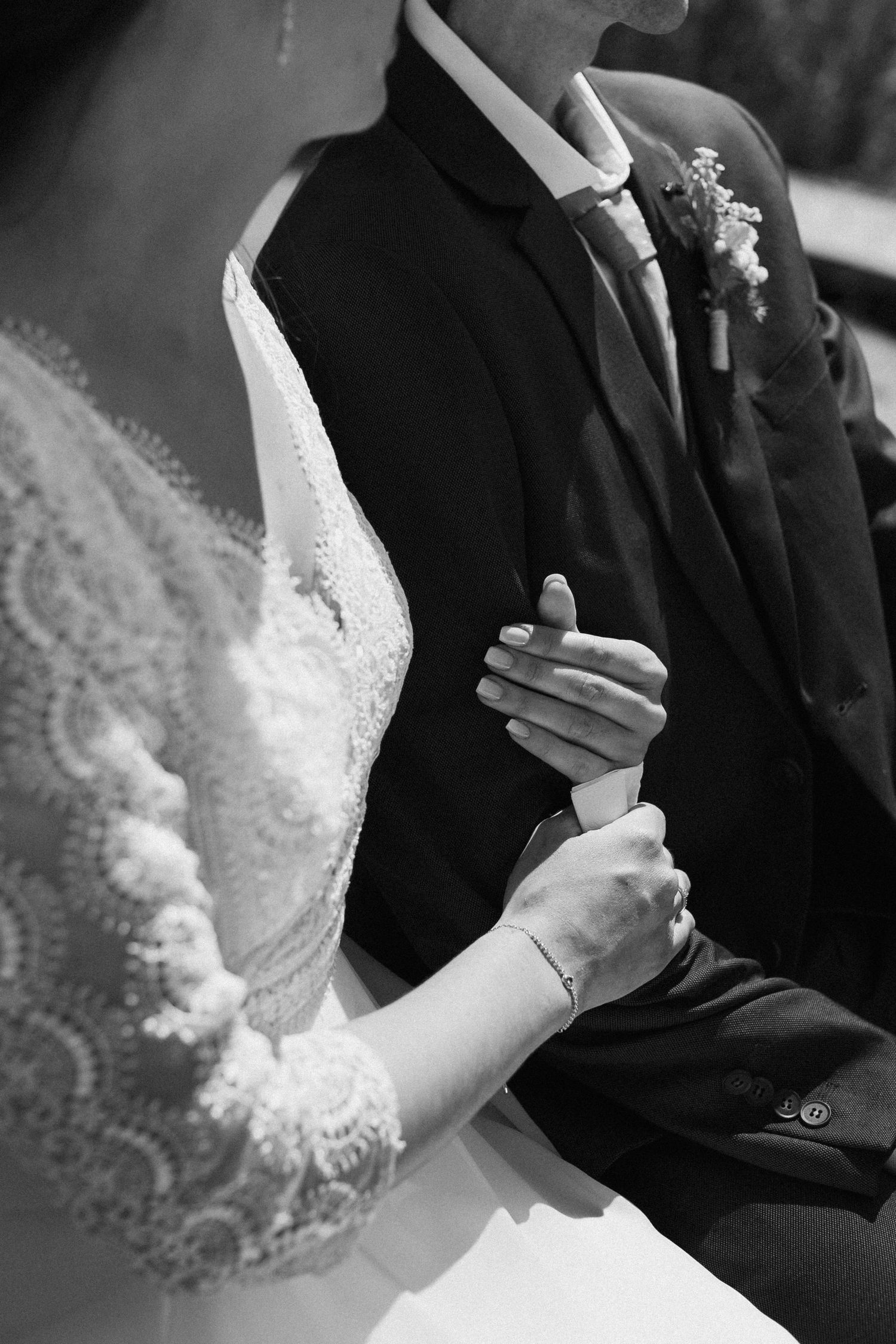 Black and white wedding photo of bride and groom holding hands, showcasing lace dress and boutonniere suit.