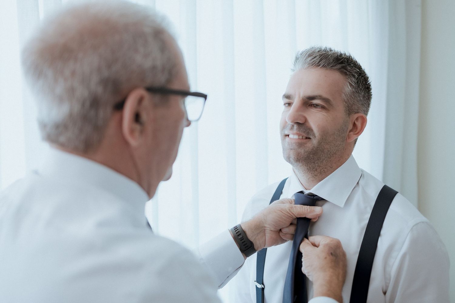 Older man adjusting tie of smiling middle-aged man wearing suspenders in bright room.