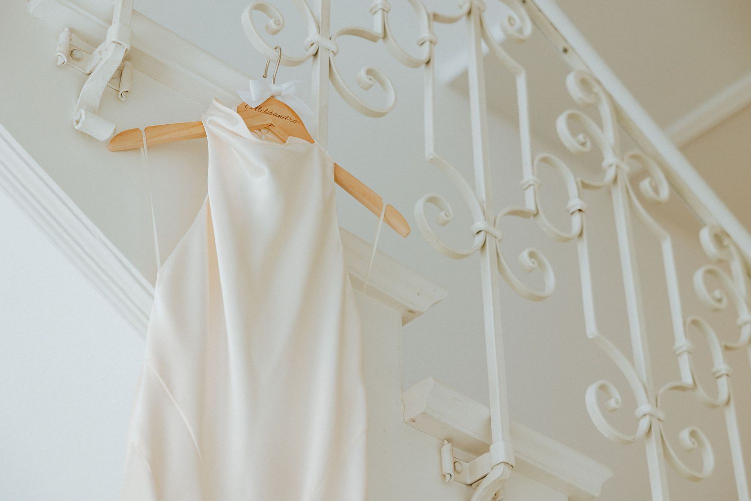 Elegant white wedding dress hanging on wooden hangers from ornate white staircase railing in bright interior.