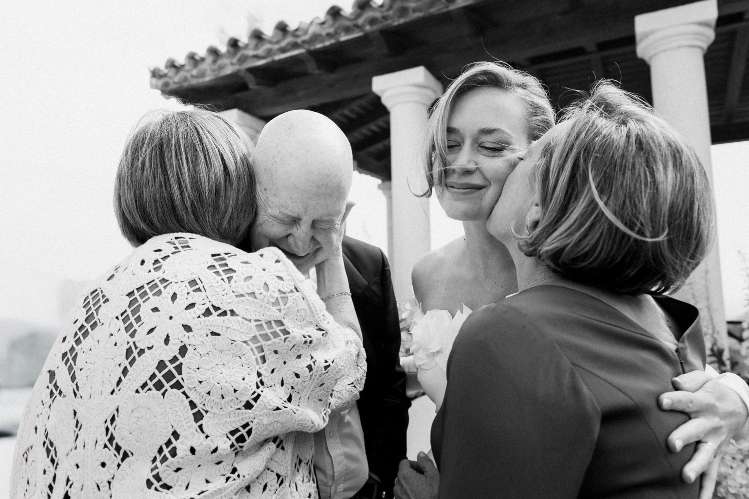 Black and white wedding photo of bride sharing emotional hugs with family members outdoors.
